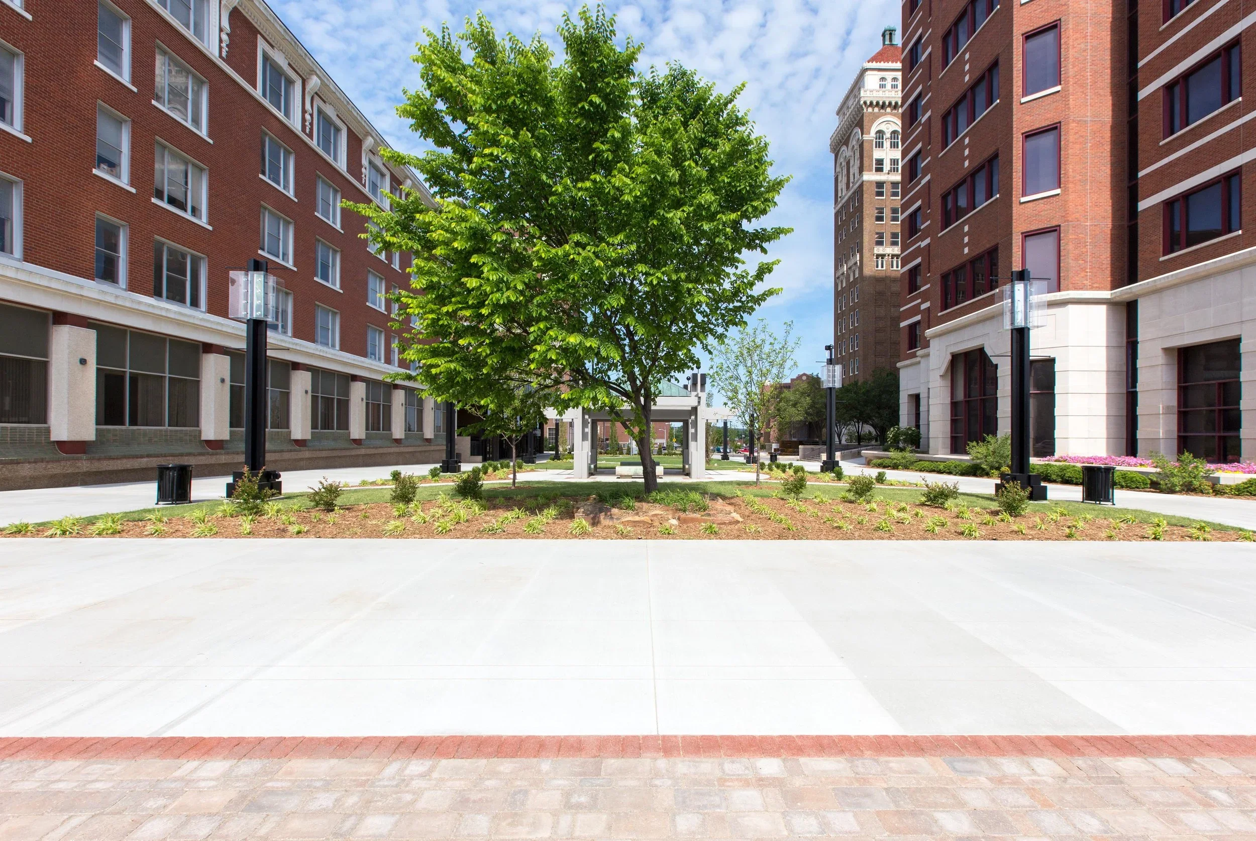 A city plaza with a central green tree, surrounding landscaped flower beds, and walking paths, flanked by tall brick apartment buildings under a partly cloudy sky.