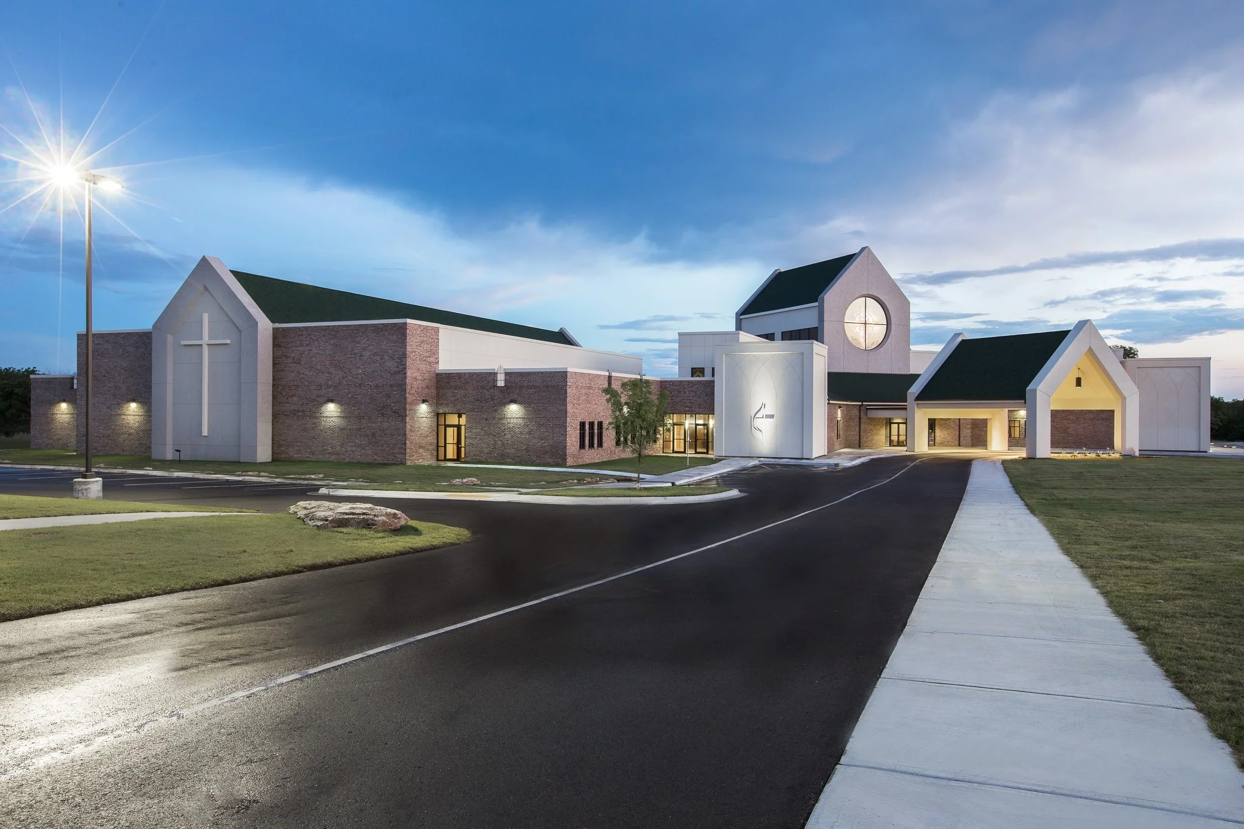 Modern church building with brick and white exterior, large cross, and circular window, illuminated at dusk with surrounding parking lot and sidewalk.