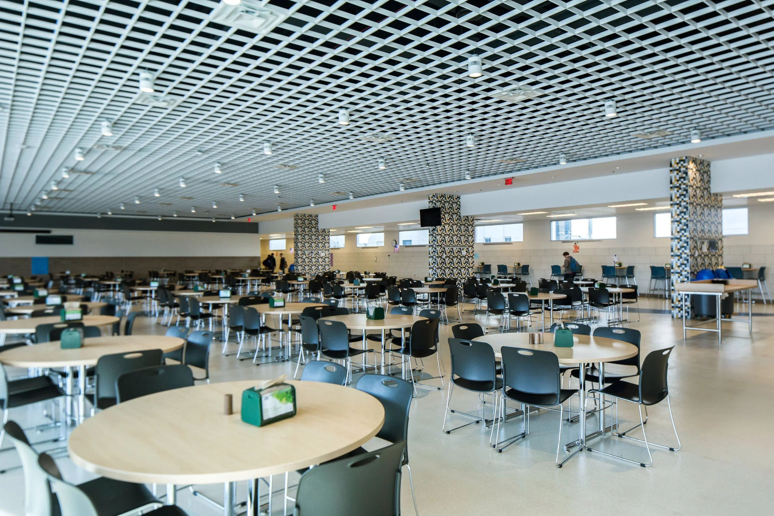 Empty cafeteria with round tables, black chairs, and condiment holders, bright windows, and patterned pillar columns.