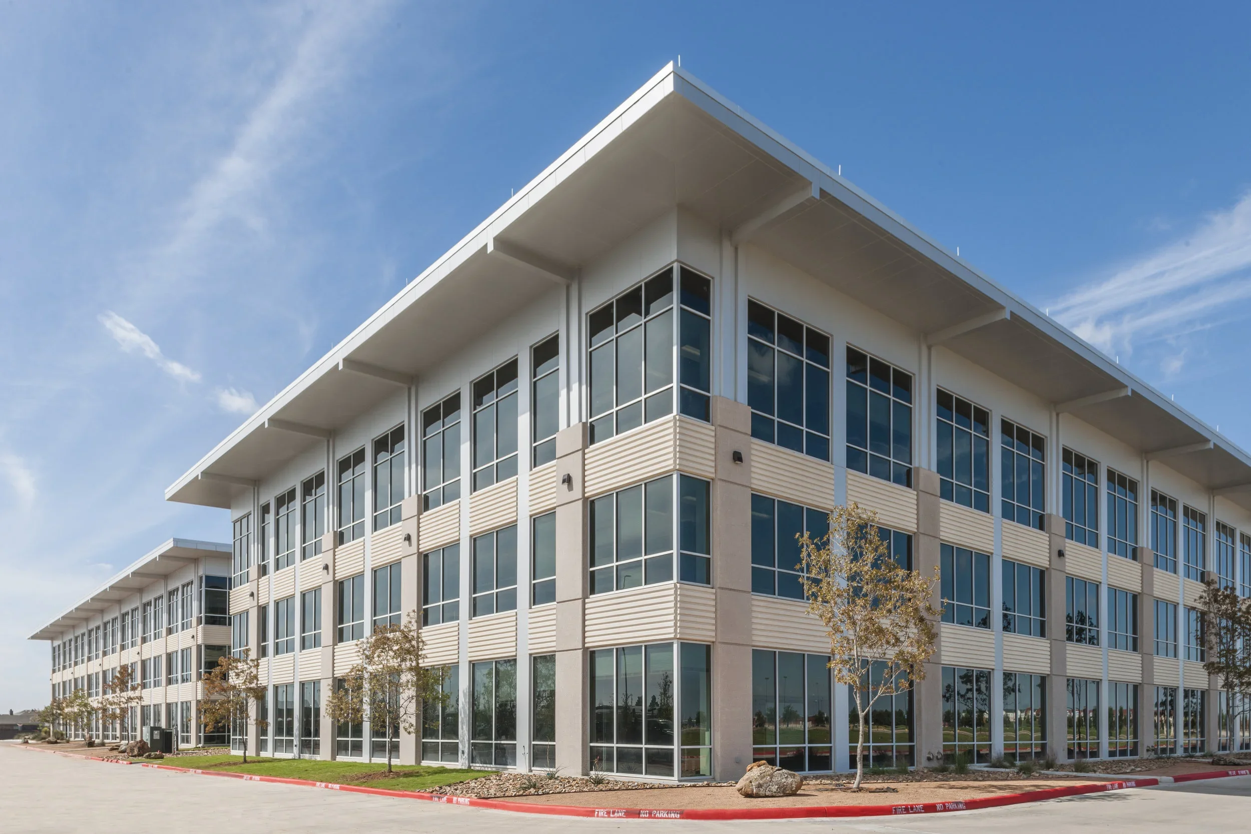 Modern 3-story office building with large glass windows, white exterior, and small trees along the sidewalk under a blue sky.
