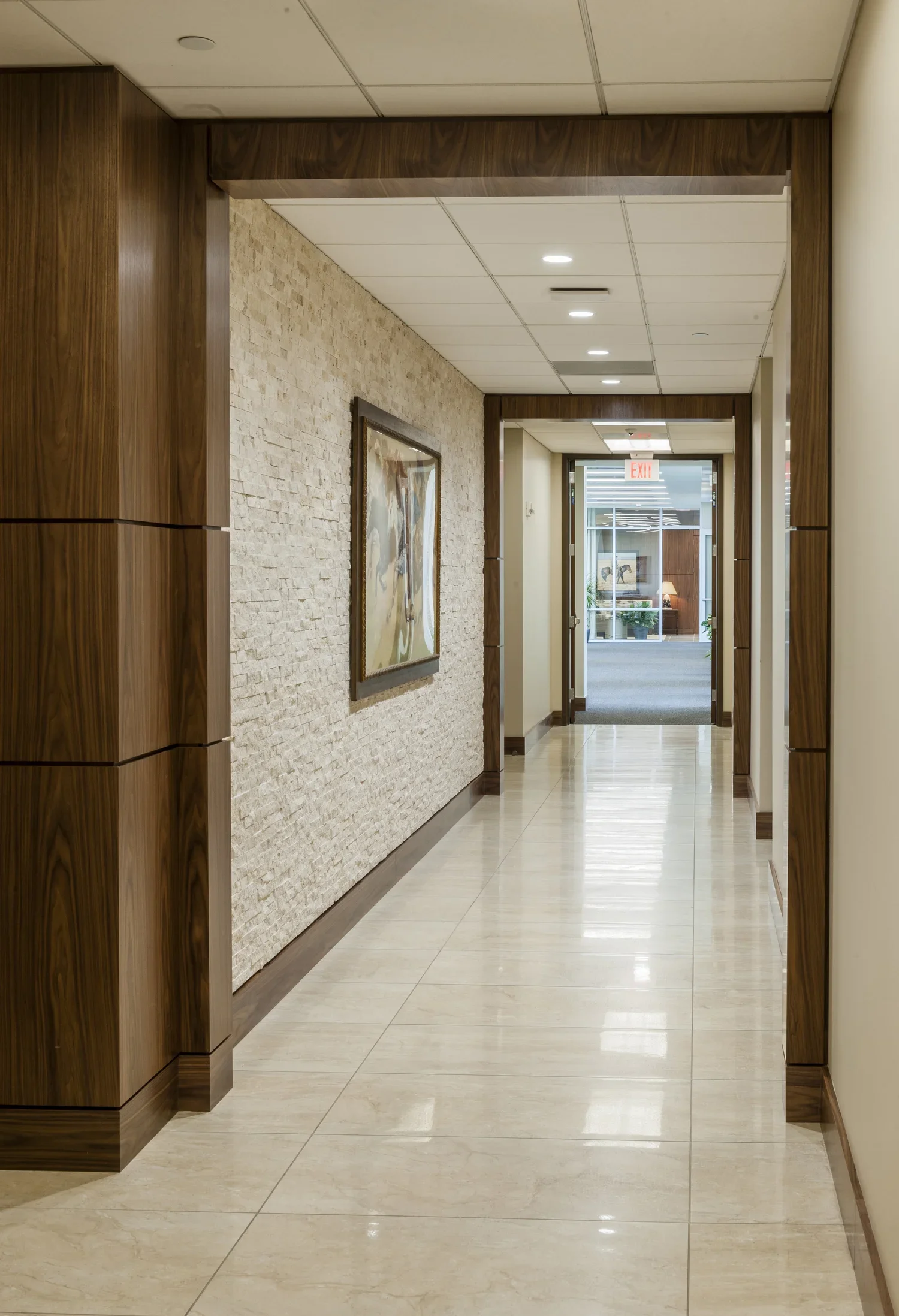 Empty corridor in an office building with a textured stone wall, framed artwork, wooden paneling, ceiling lights, and a glass door at the end leading outside.