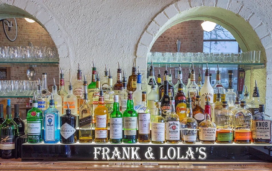 An array of various liquor bottles on a bar counter in front of a black sign that reads 'FRANK & LOLA'S'. Behind the bottles, shelves hold glasses, with arched brick and white wall backgrounds.