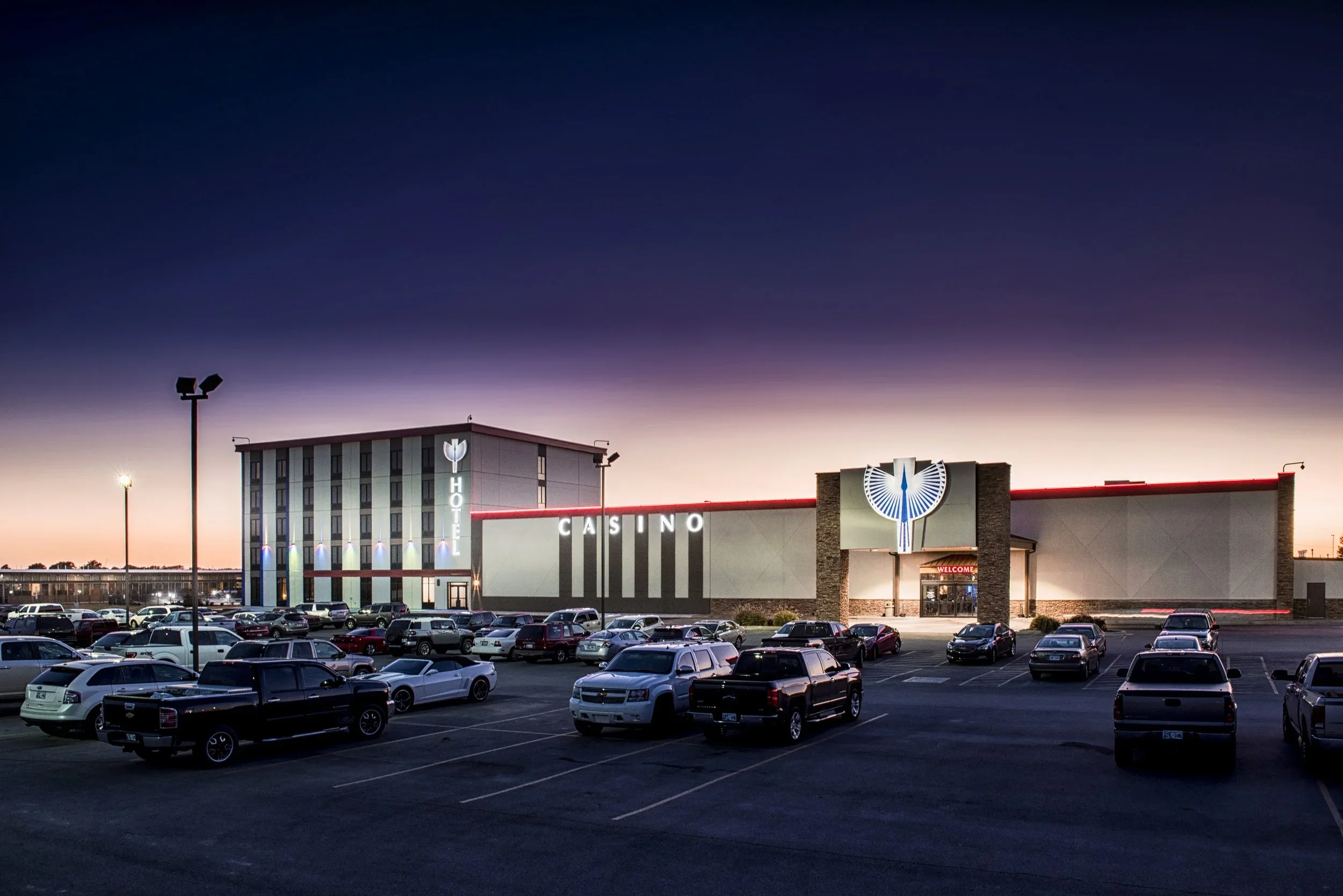 Parking lot in front of a casino and hotel building during twilight with parked cars and lit signage.