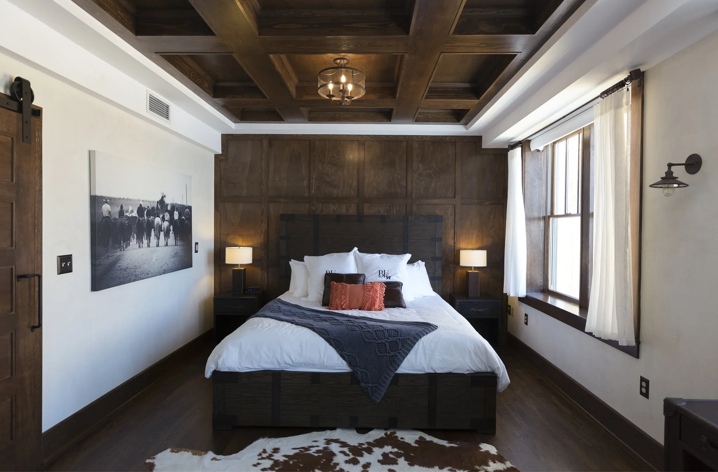 A bedroom with a large bed with white sheets, decorative pillows, and a dark blanket, flanked by two bedside lamps, a cowhide rug on the wooden floor, and wooden wall paneling behind the bed. There are windows with white curtains on the right side, and a black and white picture of cowboys on horseback on the left wall.