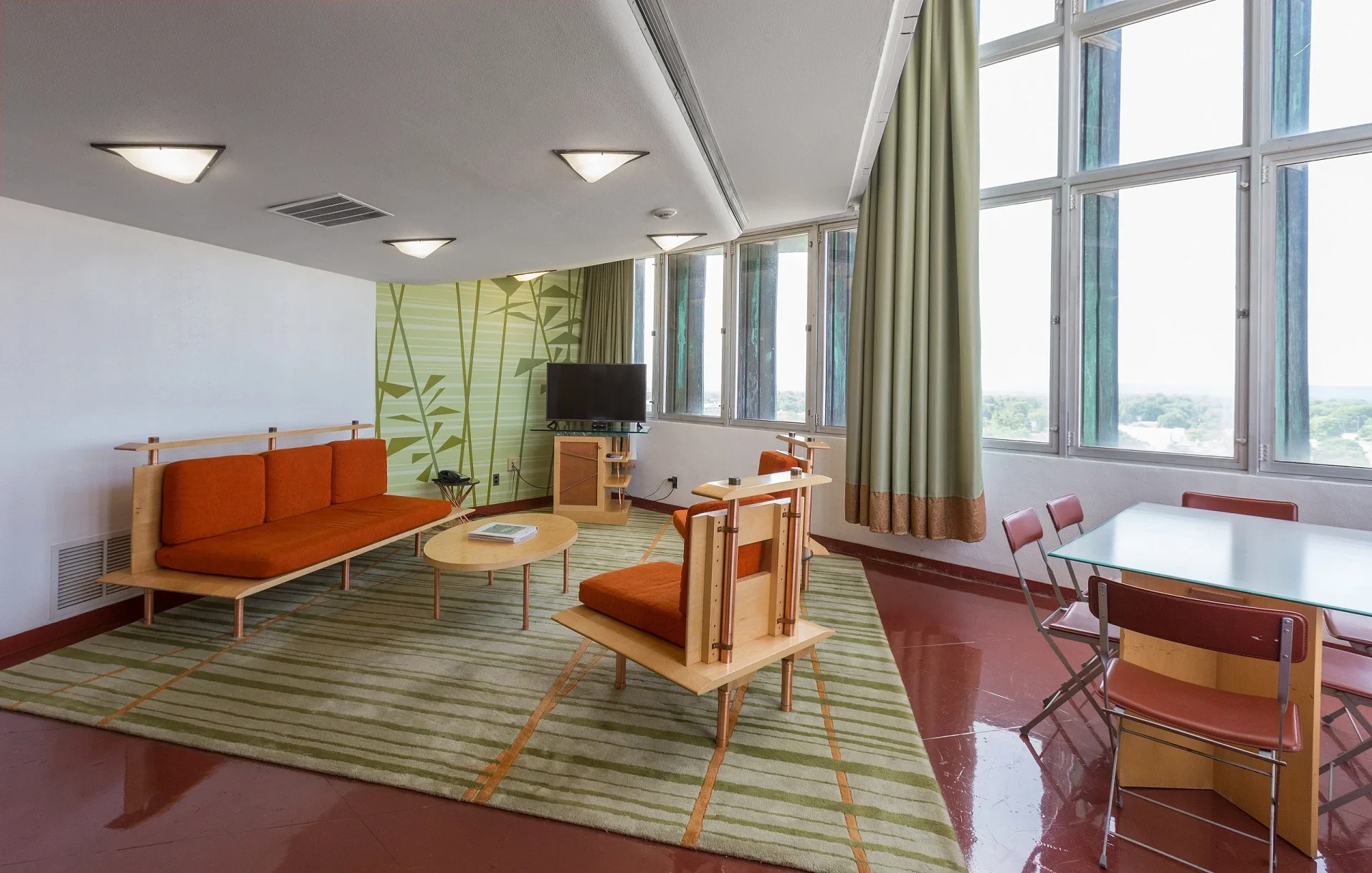 A furnished living room with a green and white patterned wall, large windows with green curtains, and orange furniture including a couch, chairs, and a TV stand. There is a glass dining table with red chairs and a patterned rug on a reddish-brown til