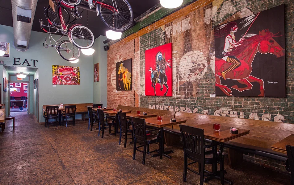 Interior of a restaurant with wooden tables and black chairs, exposed brick wall with colorful artwork, two bicycles hanging from the ceiling, and a hallway leading to another room.