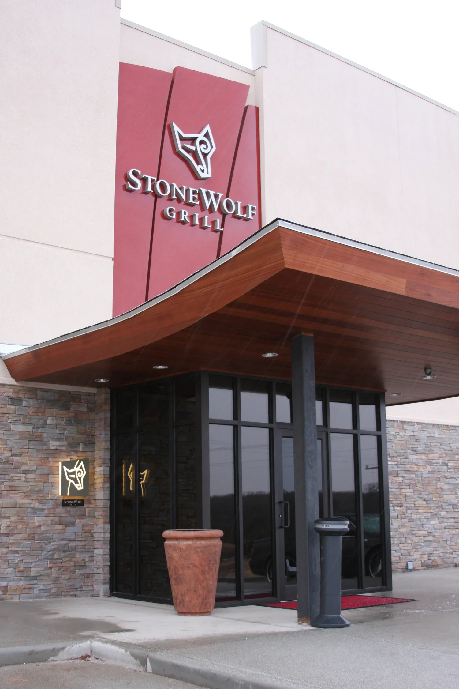 Exterior view of Stonewolf Grill restaurant, featuring a stone wall, glass entrance doors, a large planter, and a sign with a wolf logo.