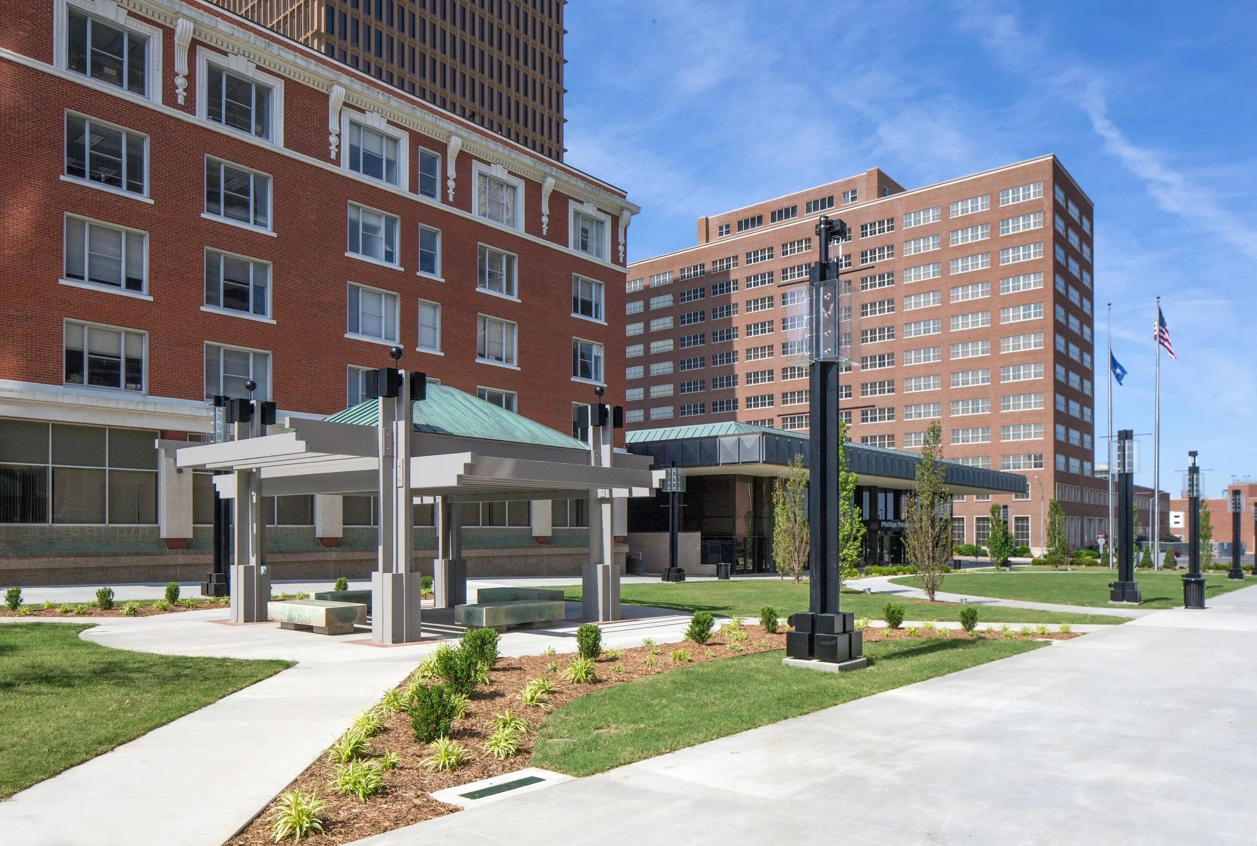 An outdoor urban setting with a small park area featuring well-maintained grass, small bushes, and pathways. There is a modern shelter with benches beneath a building with red brick walls and multiple windows. In the background, there are taller buil