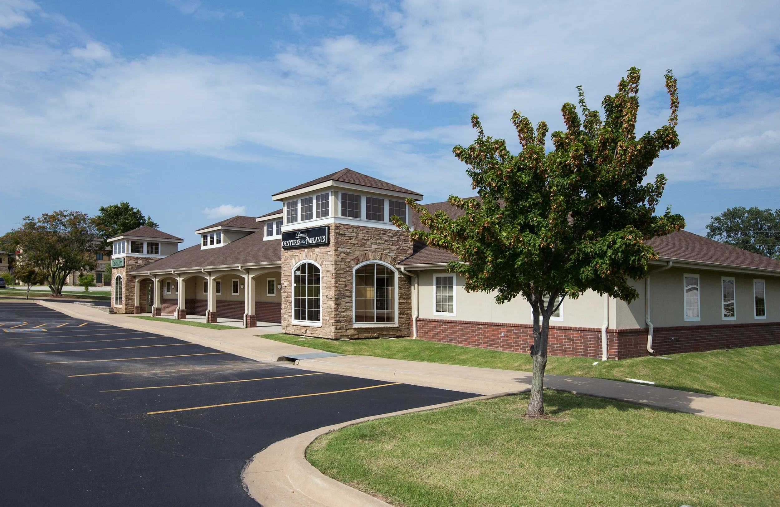A single-story commercial building with a stone and stucco facade, several arched windows, and a parking lot in front. A tree is visible in the foreground, and the sky is partly cloudy.