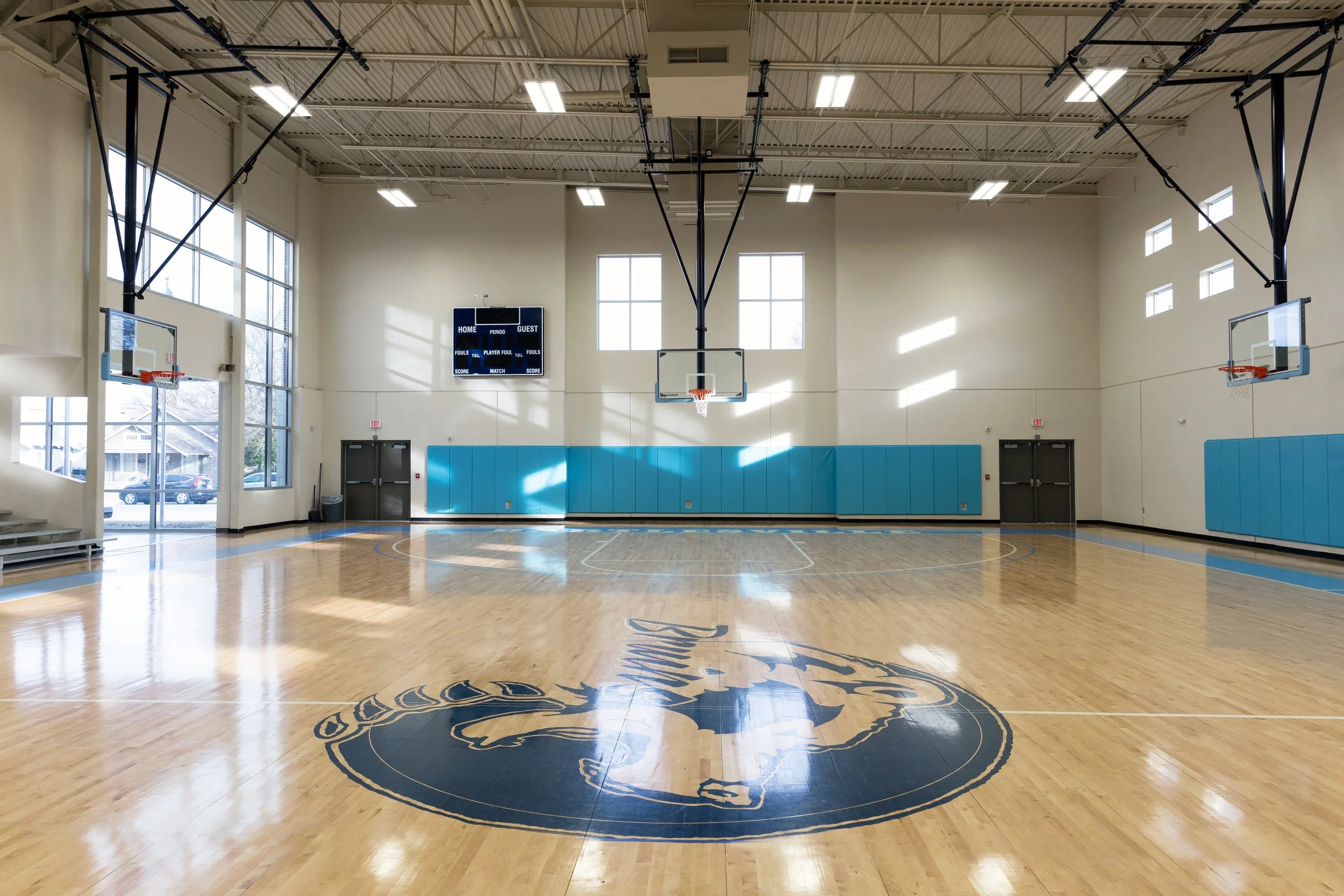 Empty indoor basketball court with four hoops, a scoreboard, and a large lion logo painted on the wooden floor.