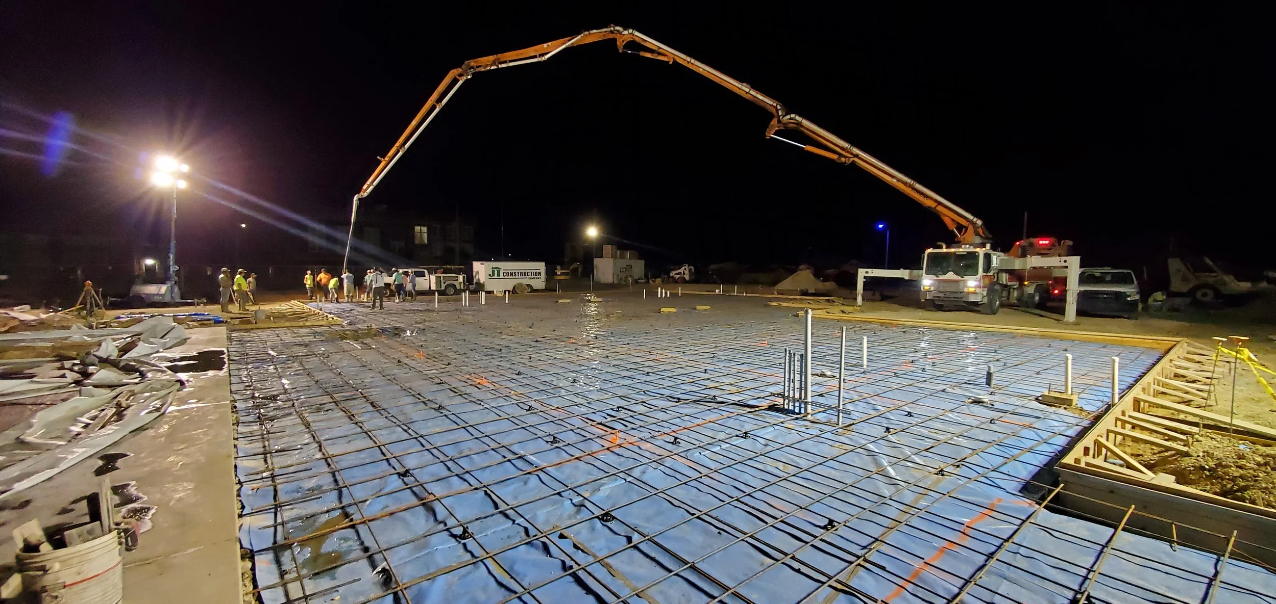 Nighttime construction site with concrete being poured over rebar reinforcement, workers, construction vehicles, and bright lights.