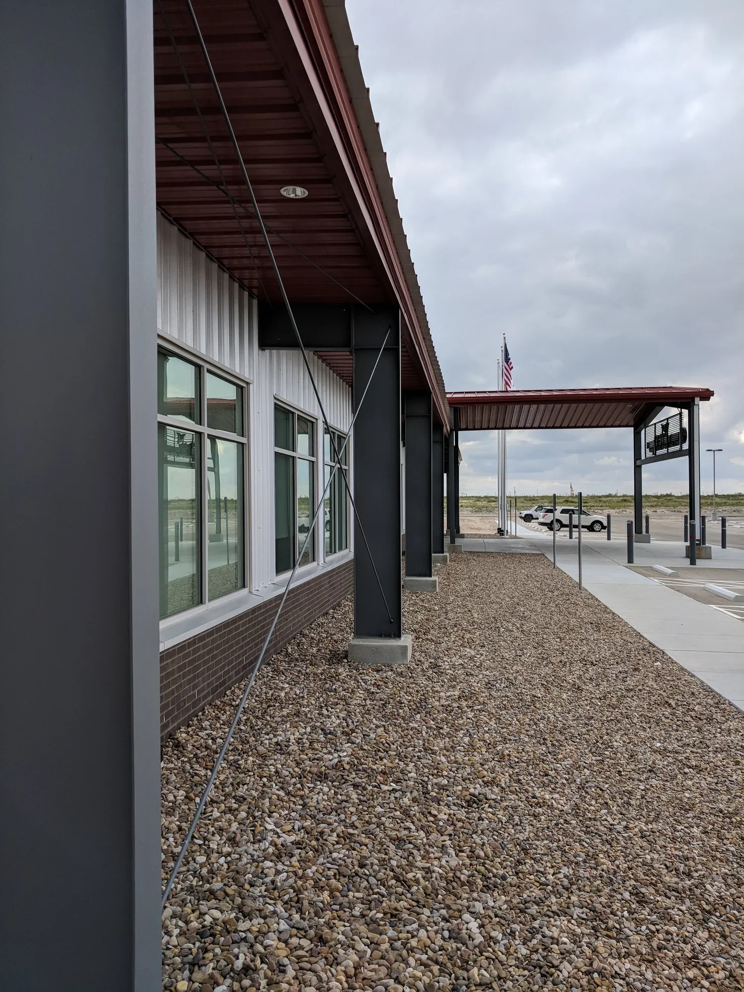 Side view of a modern commercial building with large windows, supported by steel beams, a gravel walkway, and a parking lot in the background, under cloudy skies.
