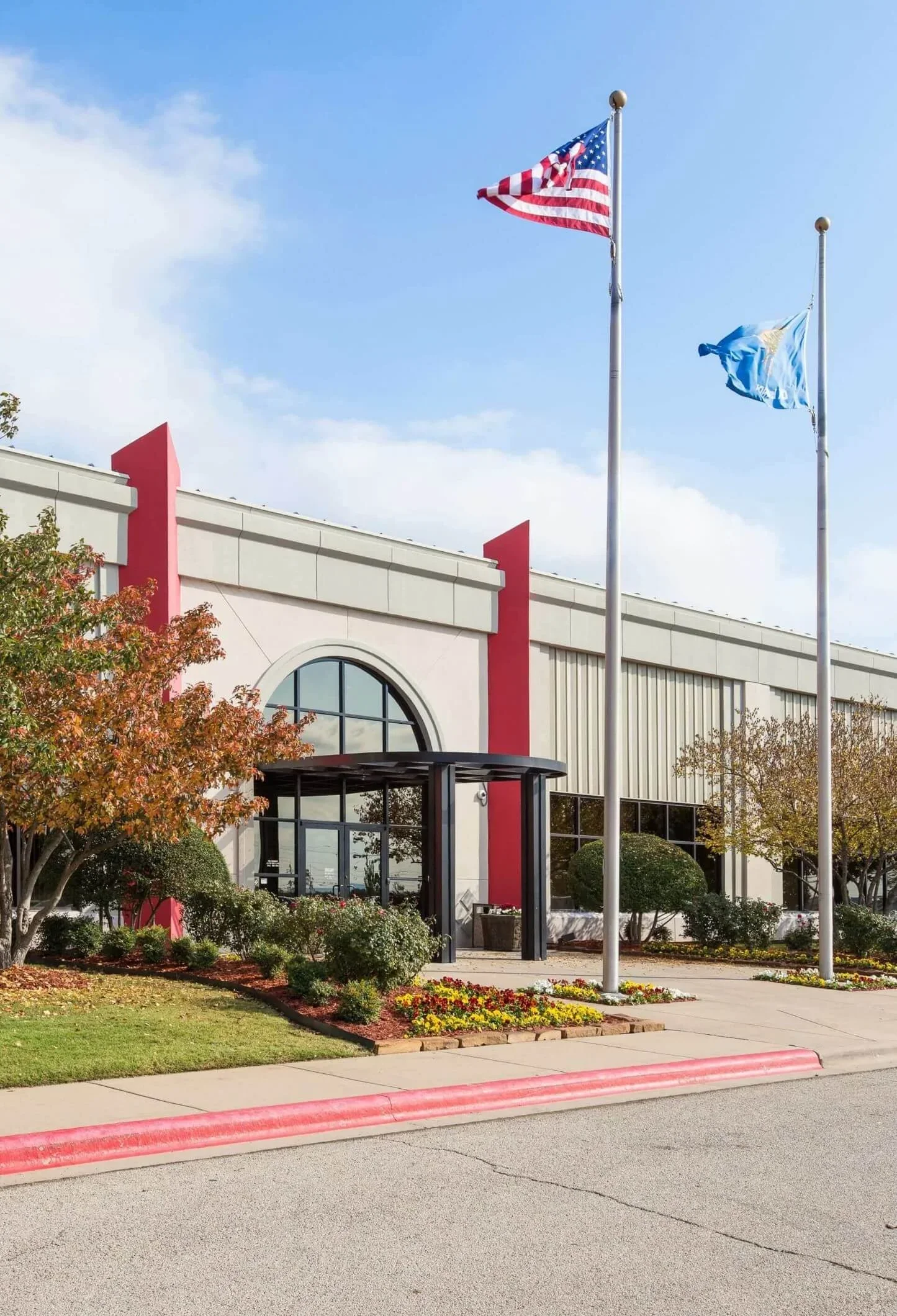 Exterior view of a commercial building with flags on flagpoles, landscaped with trees, bushes, and flowers.