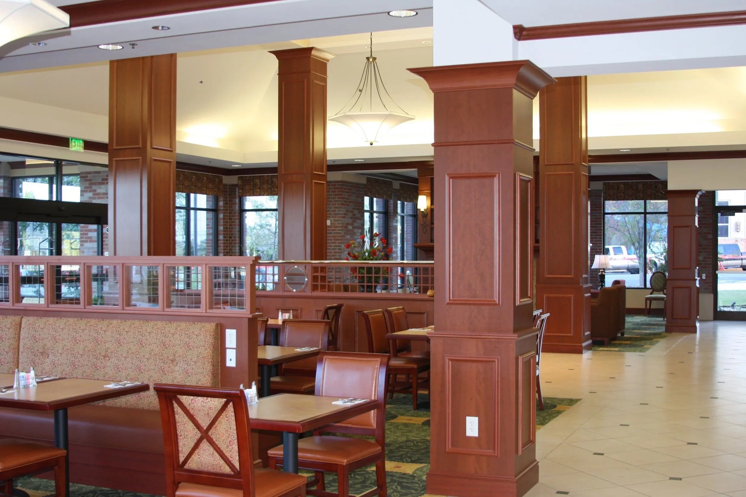 Interior of a restaurant with wooden columns, large windows, tables, and chairs, with some in booths and others free-standing, decorated with a floral arrangement and decorated with lamps and curtains.
