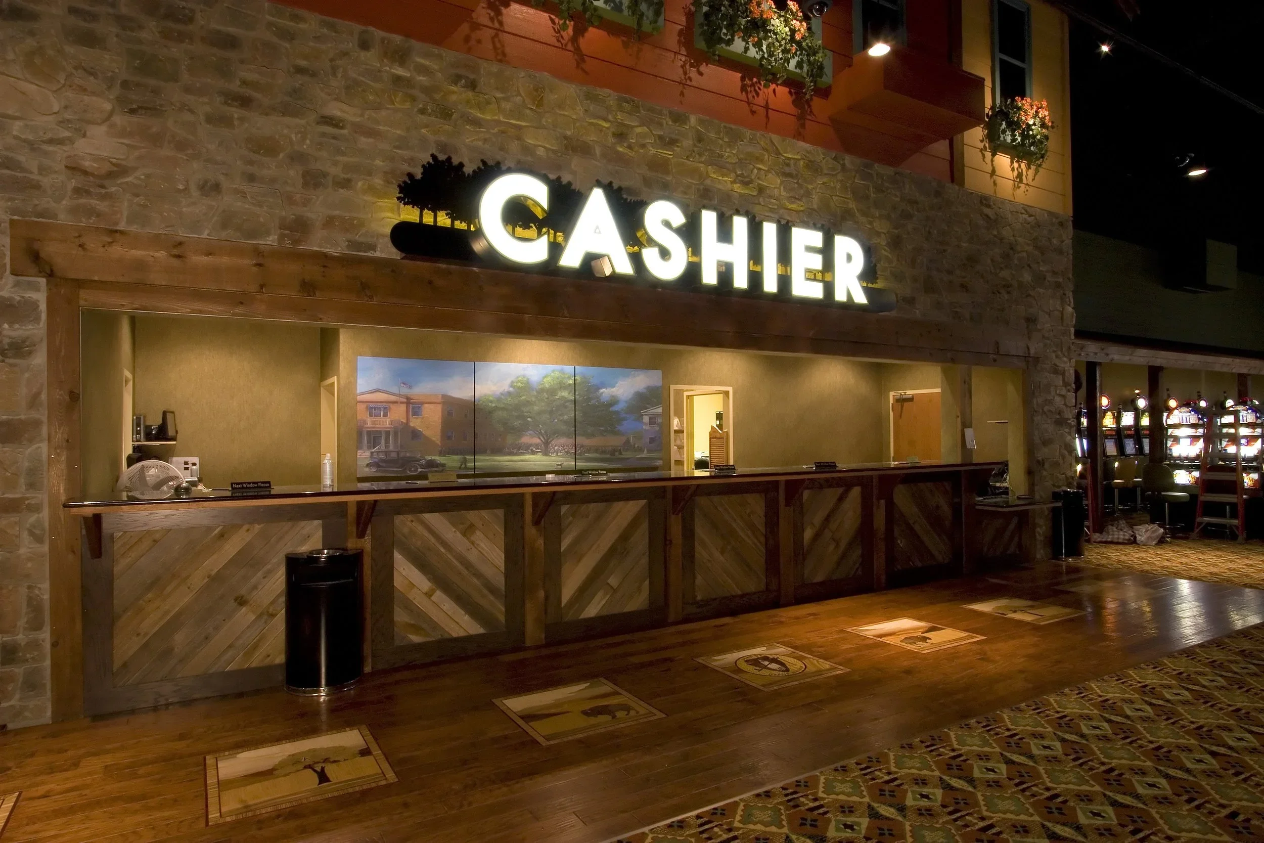 Empty casino cashier booth with illuminated 'CASHIER' sign, wooden counter, and slot machines in the background.