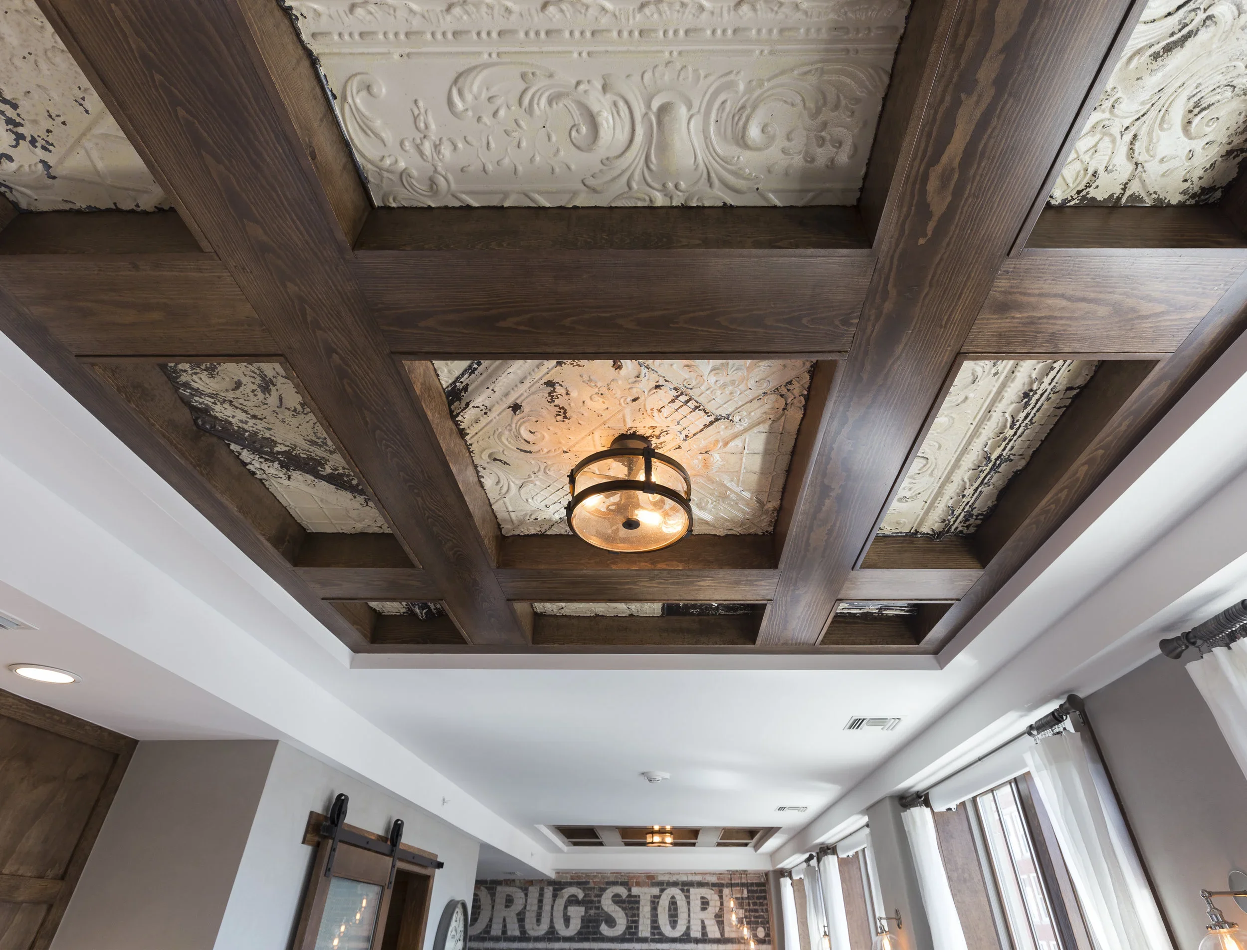 Interior view of a ceiling with a decorative wooden grid and a vintage-style ceiling light, with distressed white ceiling panels and a brick wall with a 'Drug Store' sign.