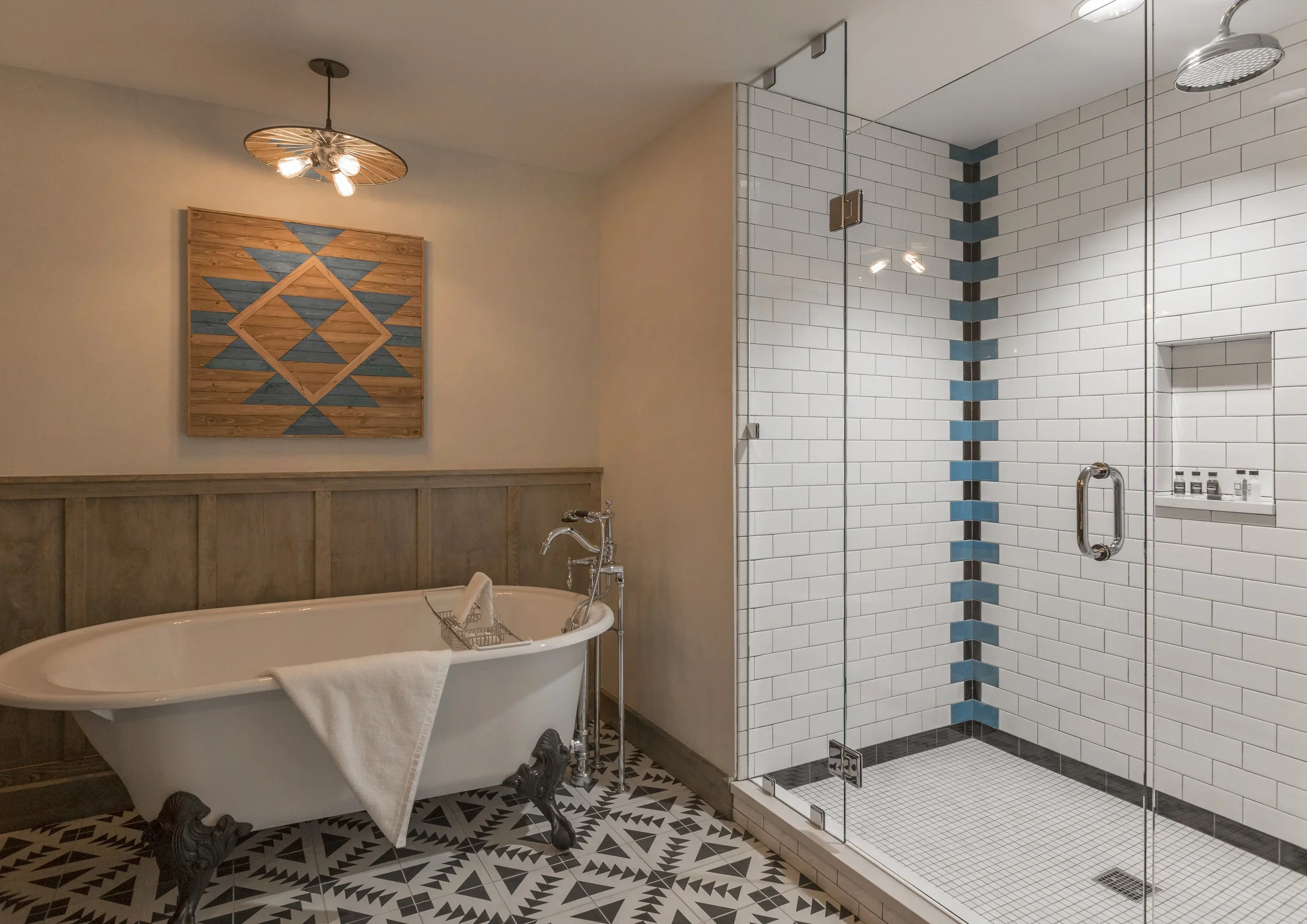 Bathroom with a clawfoot bathtub and a glass-enclosed shower, featuring white subway tiles with a pattern of alternating blue and black tiles along the corner, black and white patterned floor, wood-paneled wall behind the bathtub, and a geometric wal