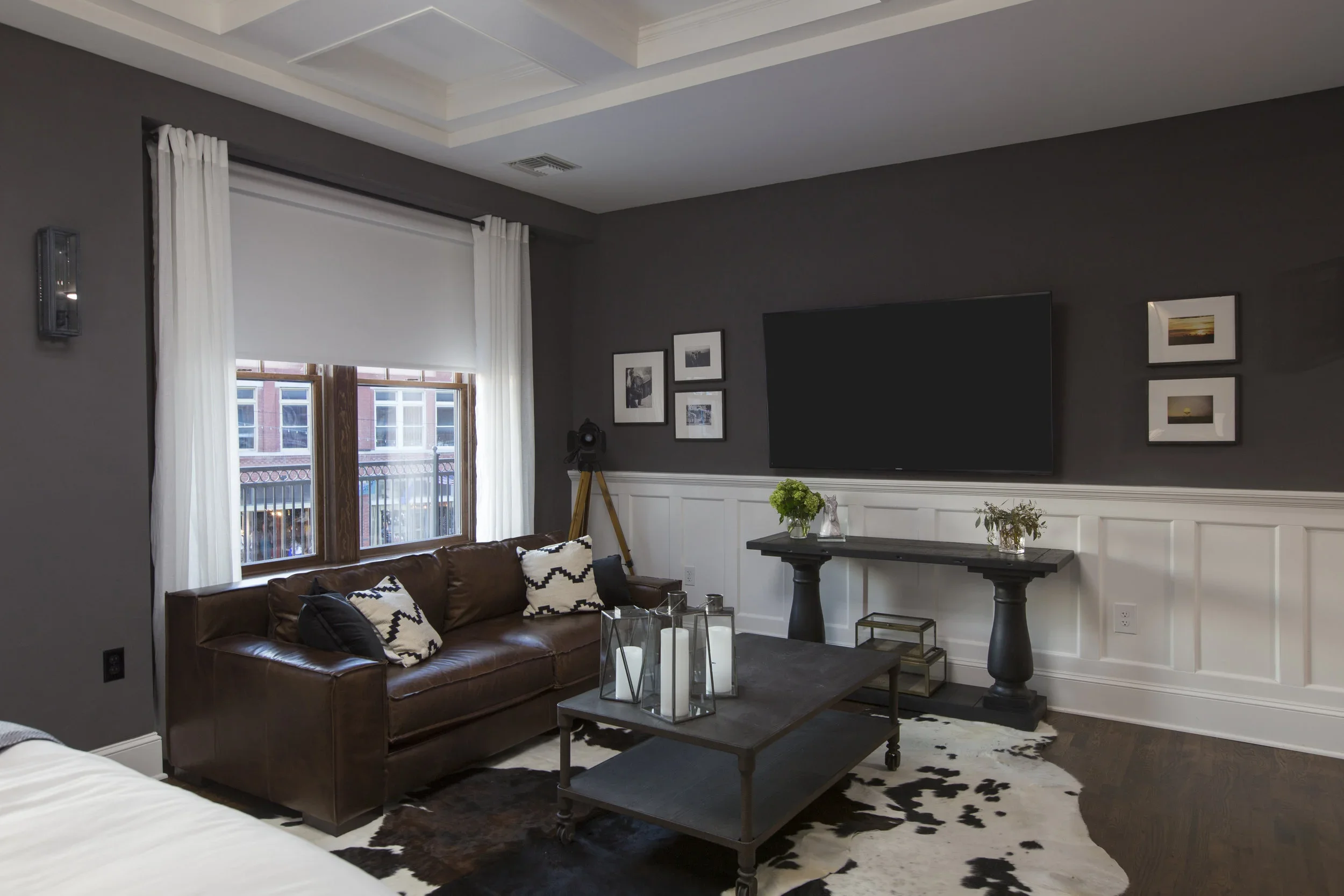 Living room with a brown leather sofa, black and white throw pillows, a window with white curtains, a dark wall with framed photos, a mounted flat-screen TV, a black console table with vase and flower decorations, a cowhide rug, and a dark wood coffe