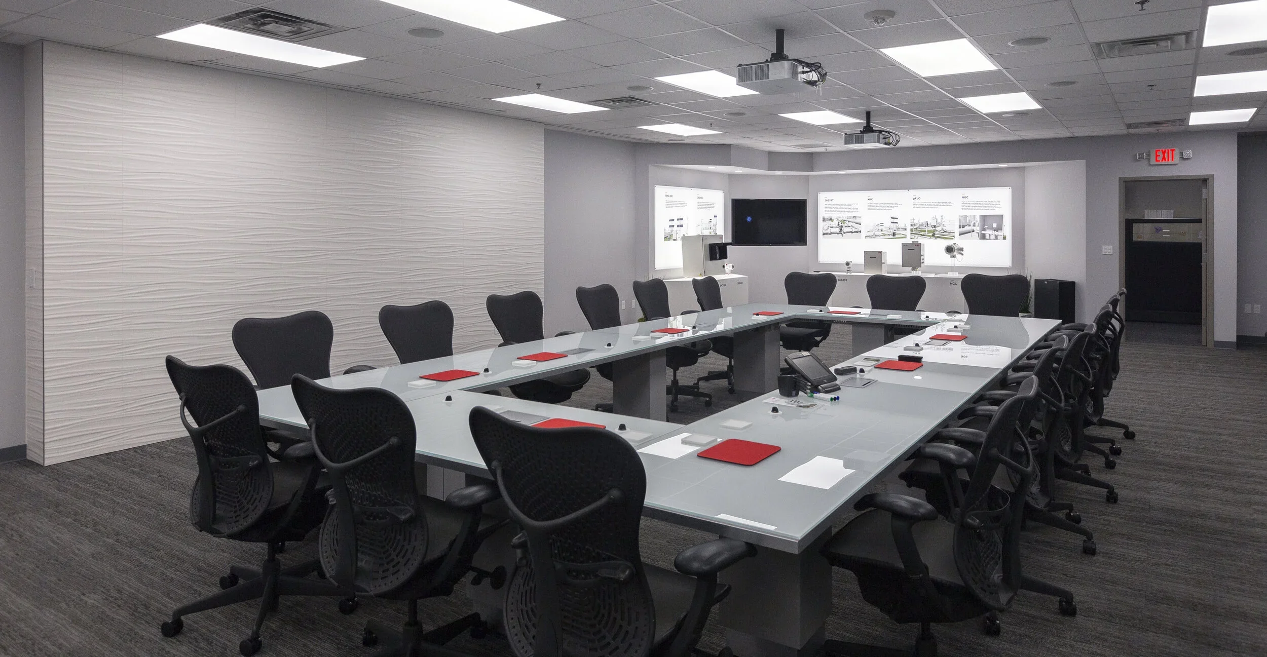 Empty conference room with a large U-shaped table, black office chairs, red notebooks, conference phone, and wall-mounted screens.