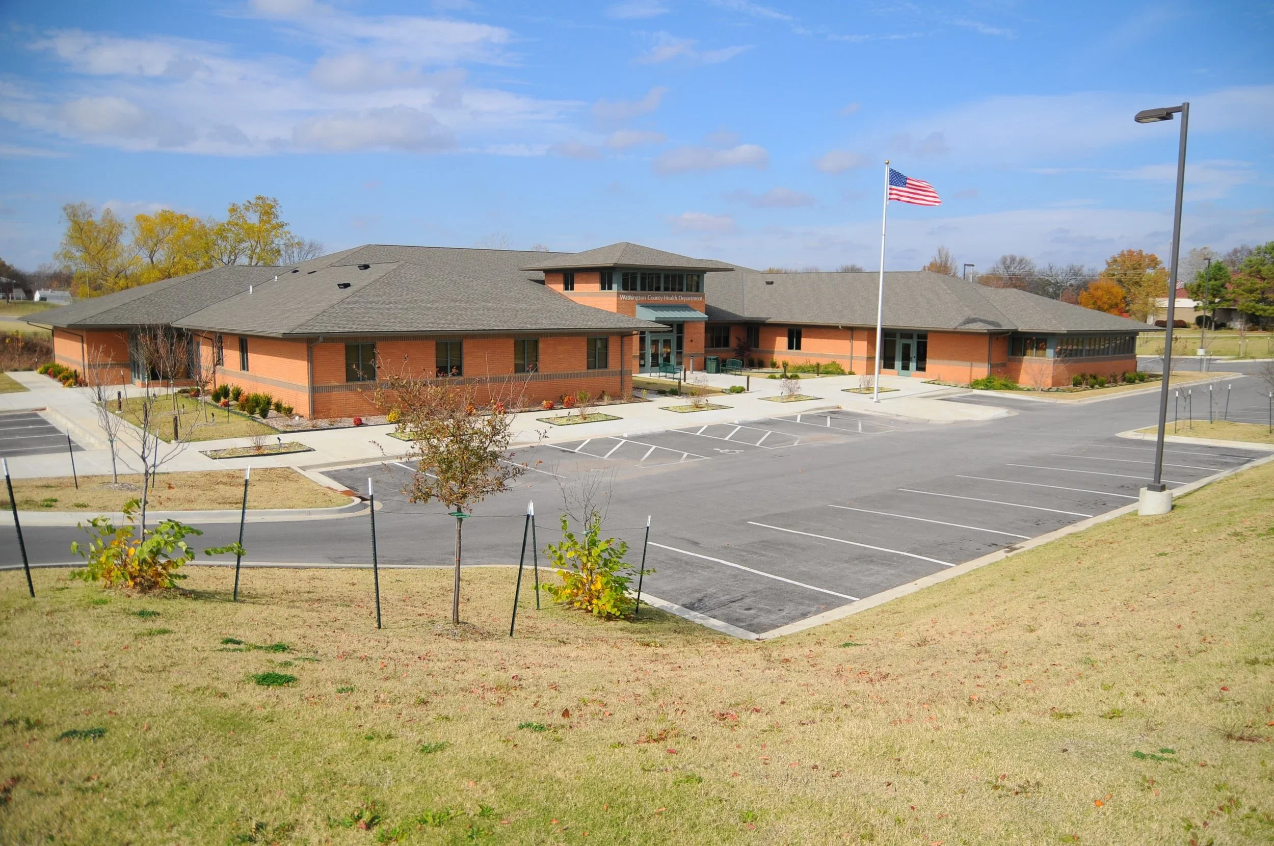 A brick building with a gray roof and a flagpole with the American flag in front, surrounded by a parking lot with empty spaces, a few young trees, and a grassy area.