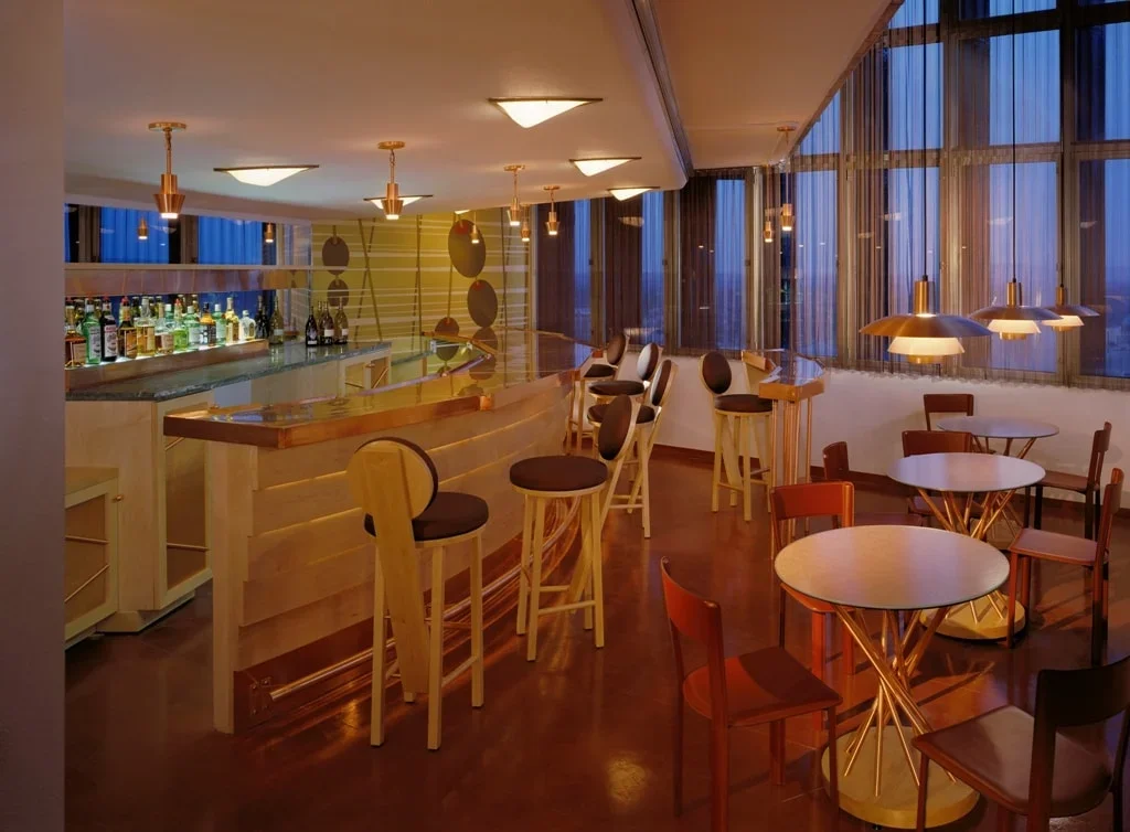 An empty bar and seating area with a curved wooden bar counter, bar stools, and round tables with chairs, illuminated by warm hanging lights and large windows with curtains.