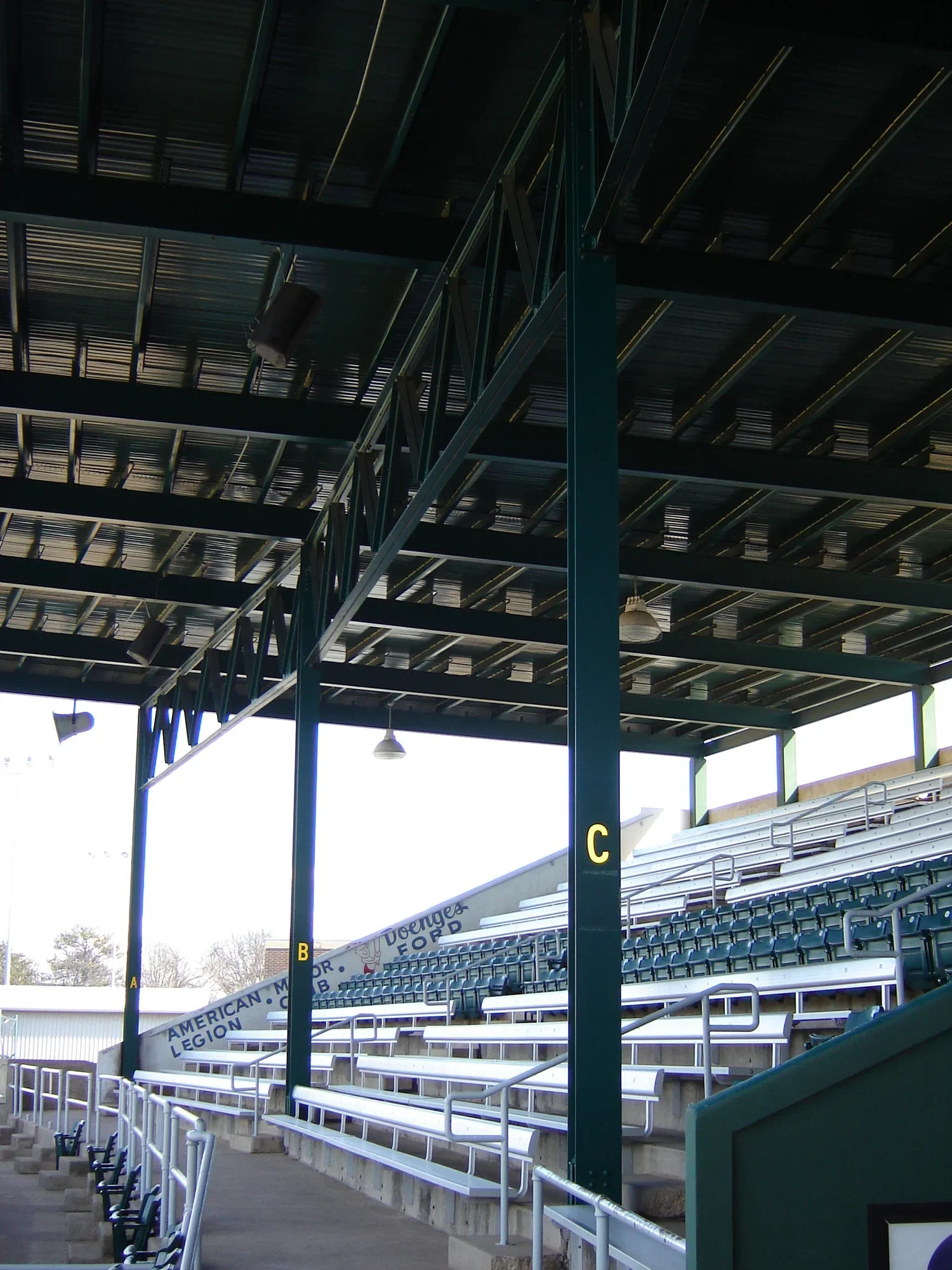 Empty bleacher seats under a metal roof at a stadium with part of a banner that reads "American Legion."
