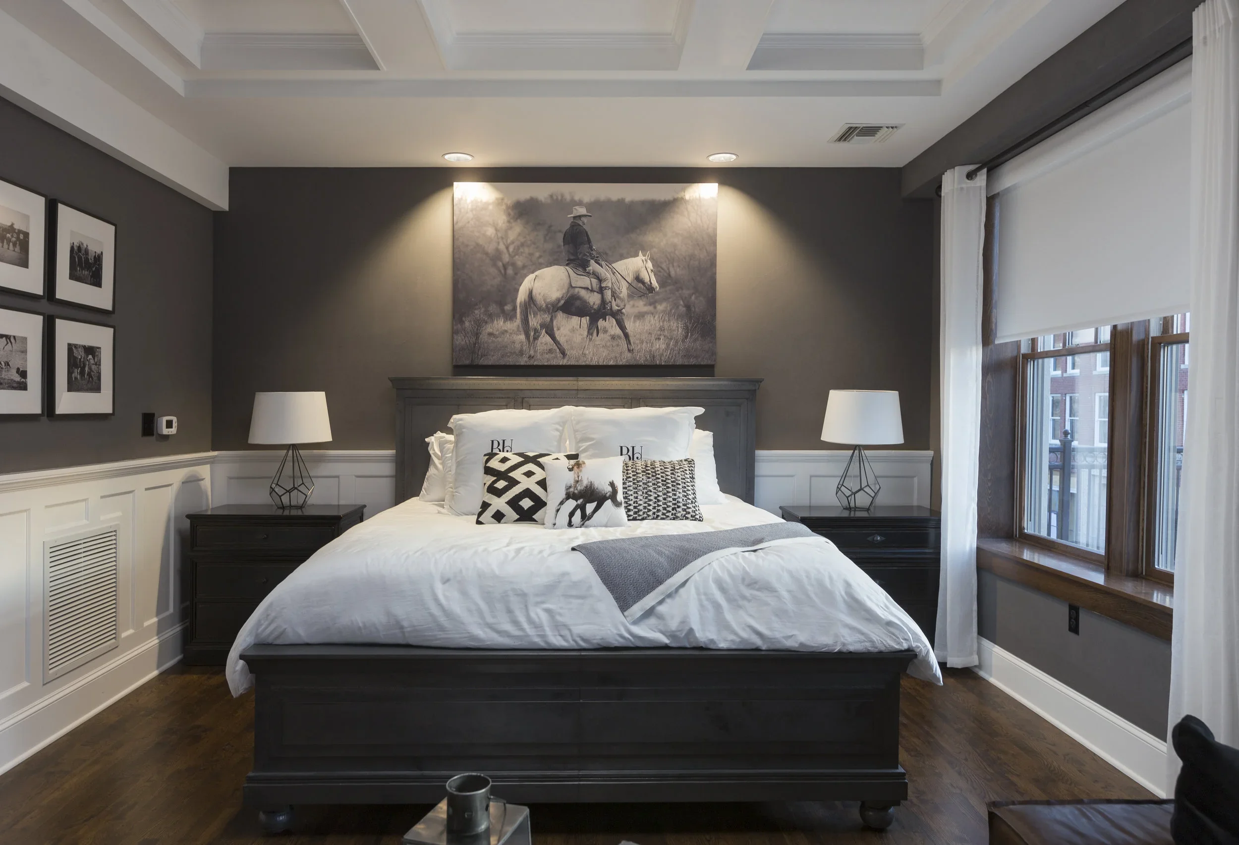 A cozy bedroom with a gray and white color scheme, featuring a large bed with white bedding, decorative pillows, and a large black-and-white photograph of a person riding a horse hanging above the headboard. There are two matching bedside tables with white lamps and framed photos on the wall to the left, a window with wooden trim and white curtains on the right, and hardwood flooring.