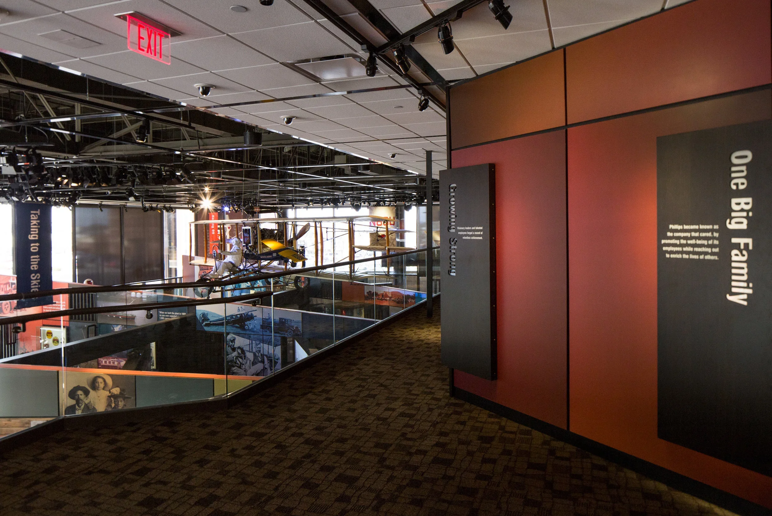 Museum exhibit with aircraft model and historical photographs, with informative panels on a red and black wall, and a staircase with glass railing.
