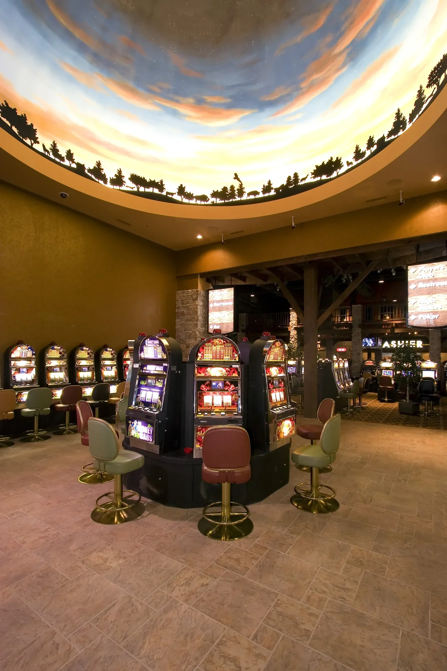 A casino with a ceiling painted to look like a sky with clouds and trees, and rows of slot machines and chairs inside.