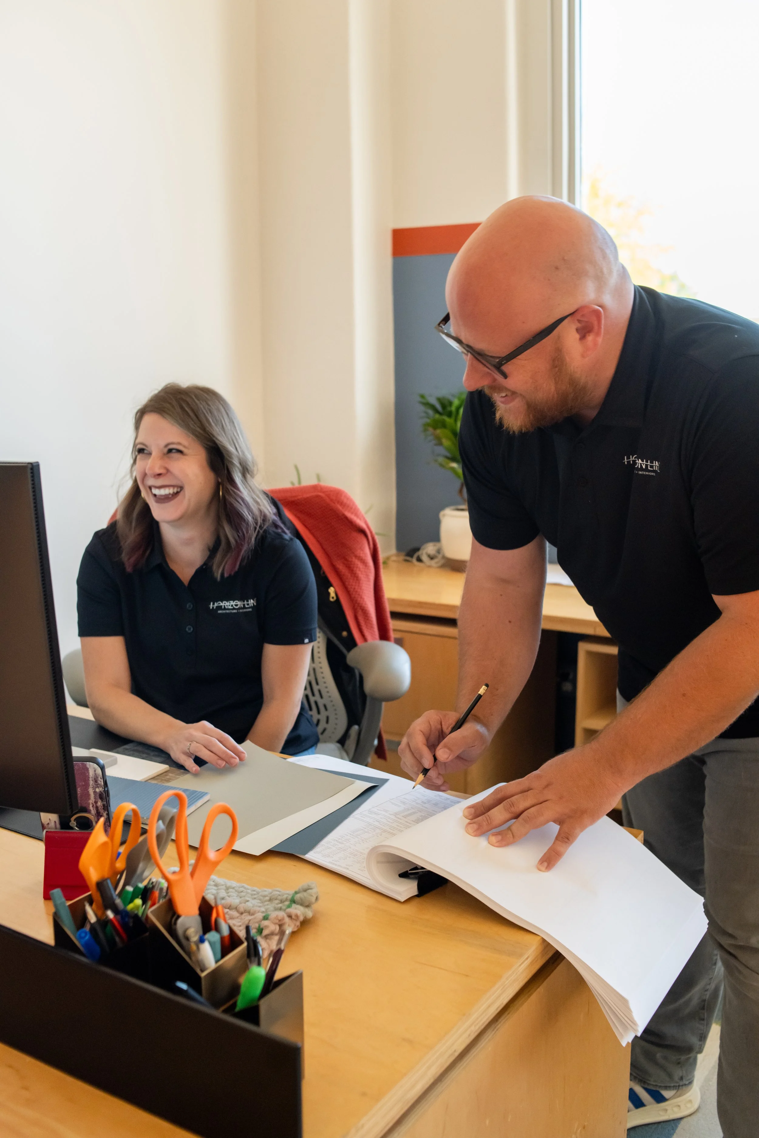 A man and woman working together at an office desk, both wearing matching black shirts with a company logo. The woman is sitting and smiling while the man is standing, writing in a large open book.
