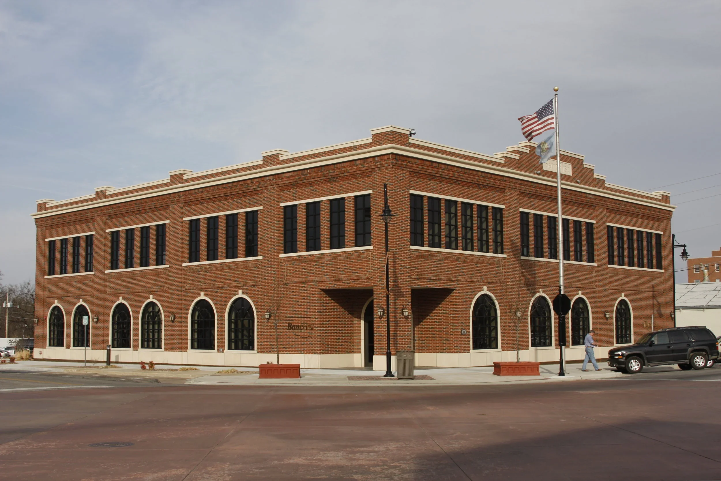 A two-story brick building with large arched windows on the ground floor and rectangular windows on the upper floor, located on a city street. An American flag and another flag are flying on a flagpole in front of the building, with a person walking on the sidewalk and a black SUV parked nearby.