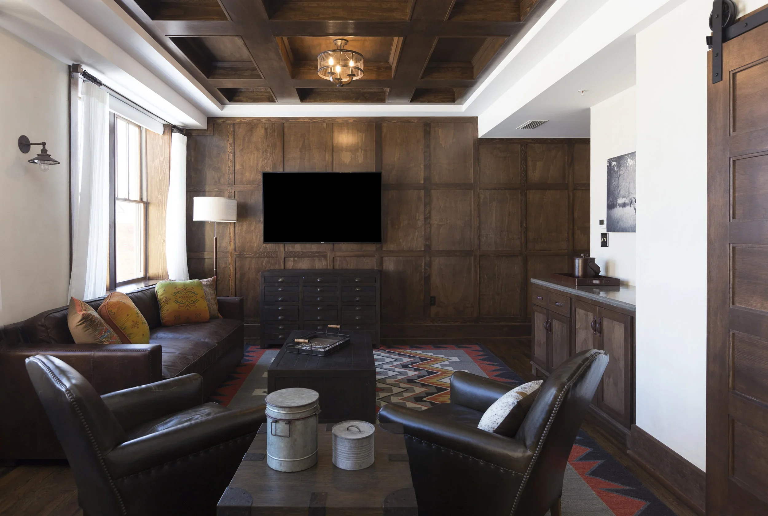 Living room with wooden paneled walls, a sofa with colorful cushions, leather armchairs, a dark wood coffee table, and a wall-mounted TV. There are windows with white curtains, a standing lamp, a sideboard, and decorative items.