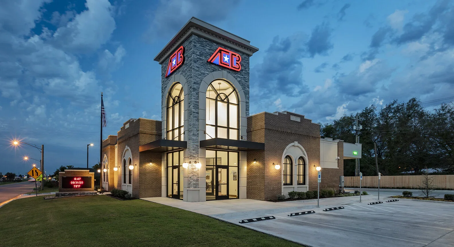 Exterior of a bank building with large windows and two illuminated signs with red and blue colors, the bank's logo contains a star. The parking lot is empty, and the sky is at dusk with some clouds.