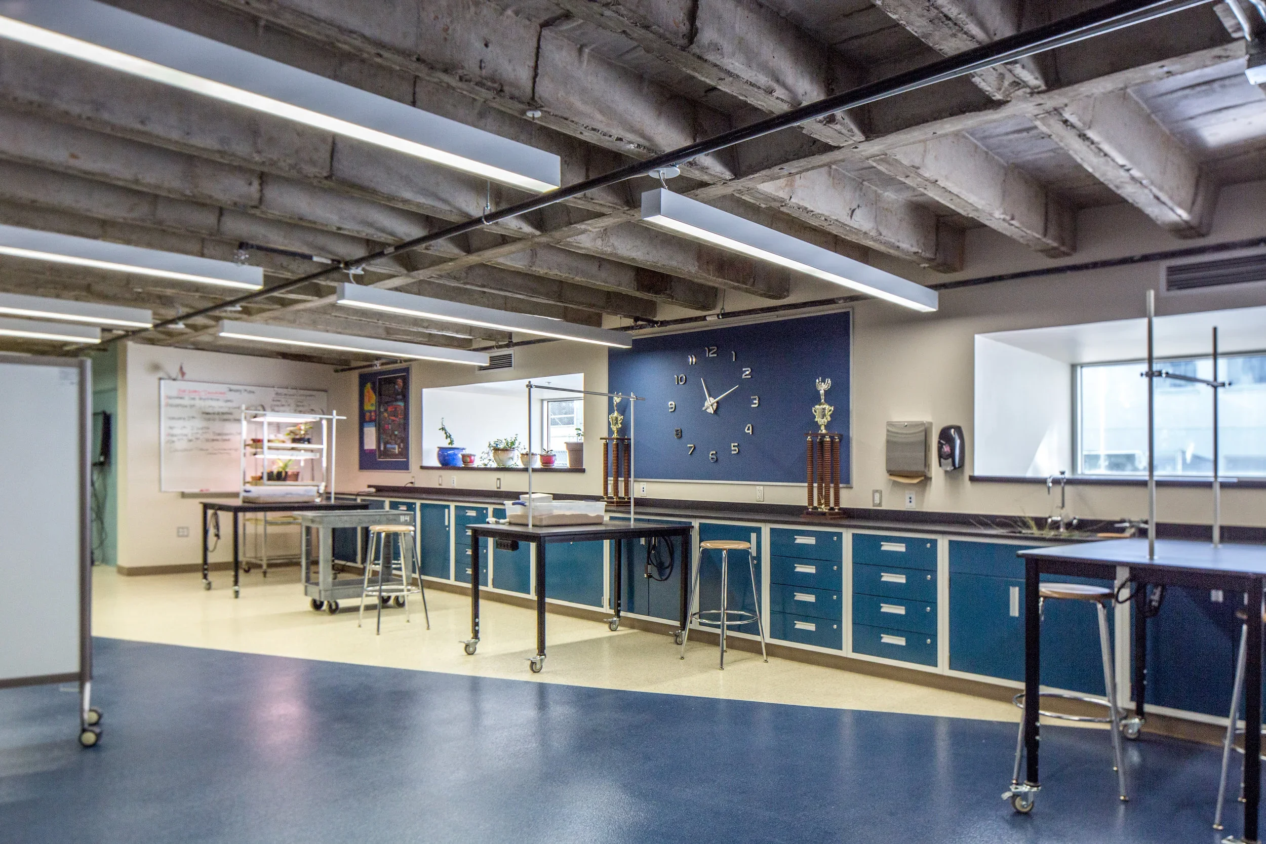 A science classroom with blue cabinets, a large wall clock, and two trophies on tall wooden stands. There are multiple tables with stools, a whiteboard, potted plants on the window sill, and a whiteboard with notes on the left side. The room has expo