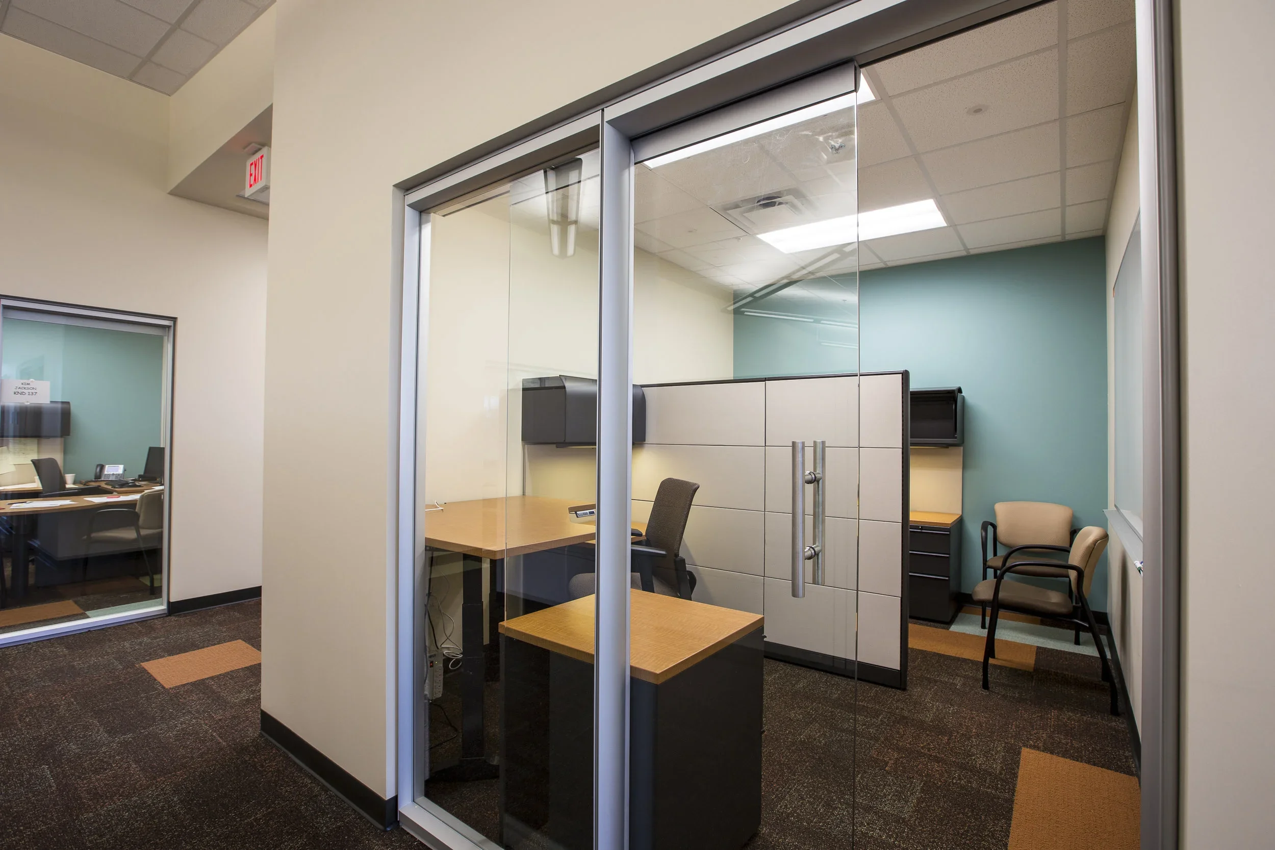 Empty office cubicle with a desk, black chair, and a partition, and a corner with two chairs and a cabinet in an office.