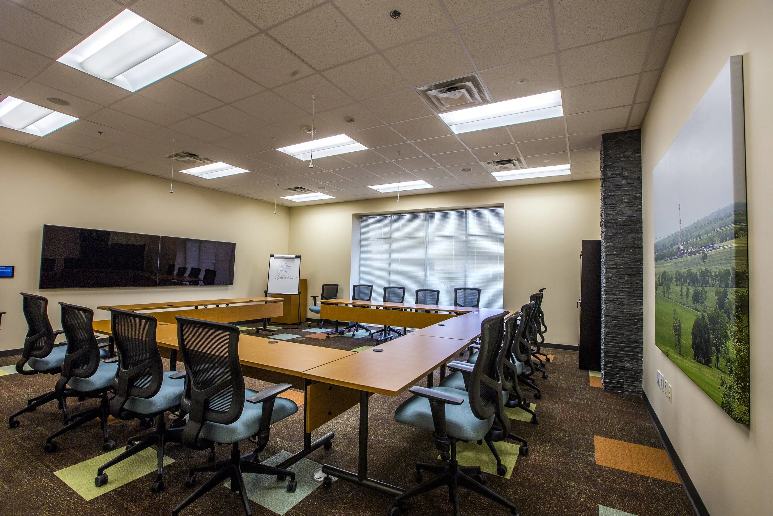 Empty conference room with U-shaped wooden tables and black office chairs, large screens on the wall, a whiteboard, and a large window with blinds.