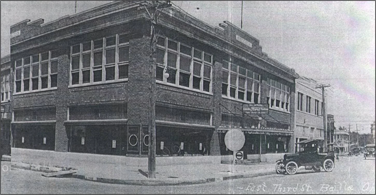 A black and white photo of a multi-story brick building on a street corner with vintage cars parked in front.