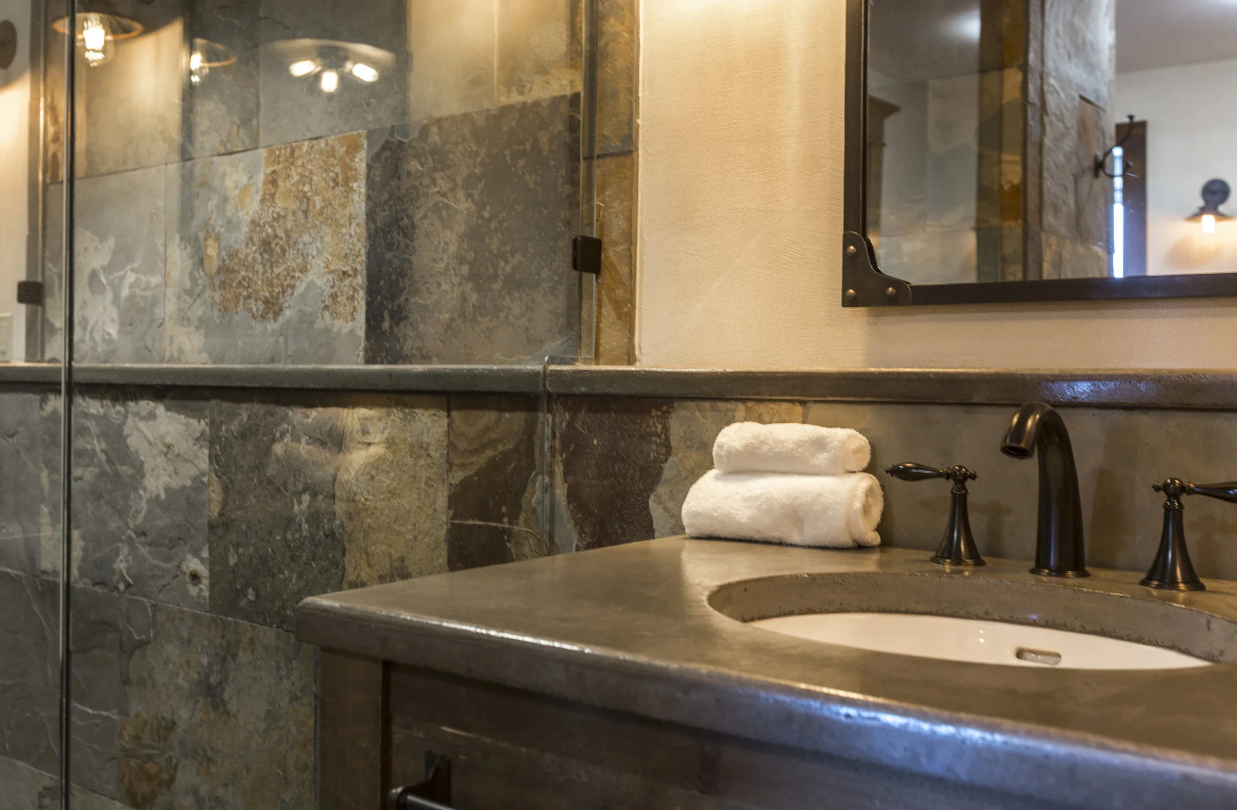 Bathroom vanity with a stone countertop, a black faucet, and three rolled white towels stacked on the counter. A mirror and tiled wall are visible in the background.