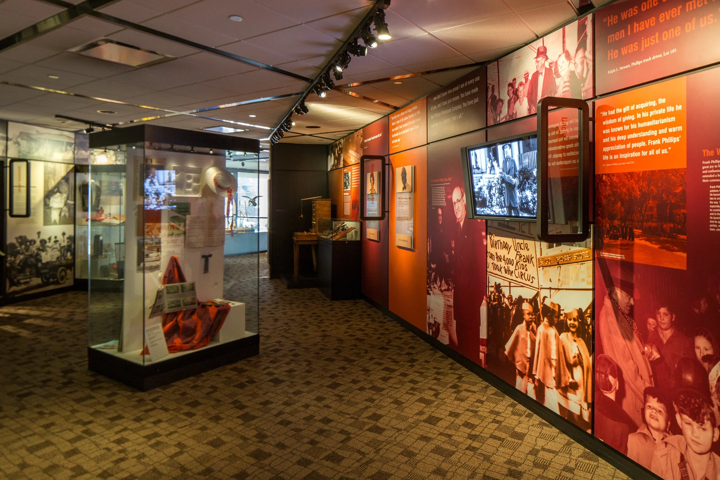 Museum exhibit with glass cases displaying historical artifacts, a red and black wall with photos, text, and framed images, some screens showing historical footage, and a carpeted floor.