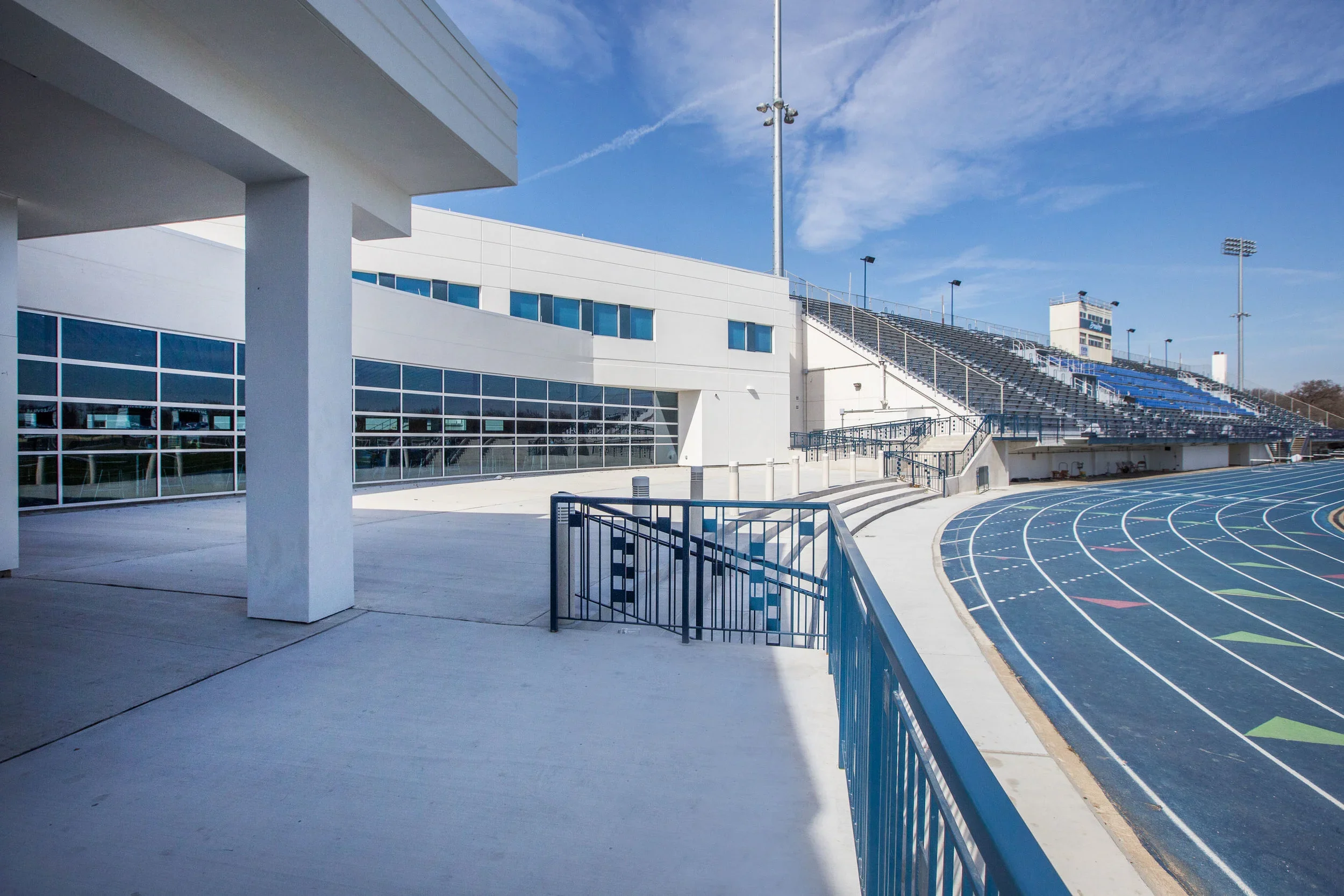 Empty school stadium with track field, bleachers, and modern white building under a blue sky.