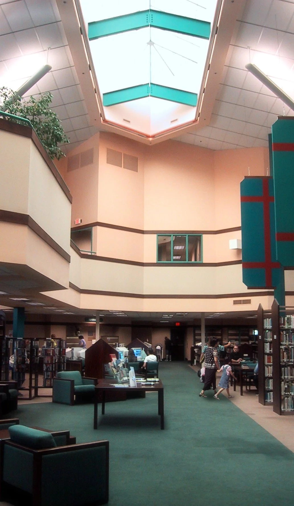 Interior of a public library with bookshelves, tables, and seating area, with a second story balcony and a large skylight ceiling.