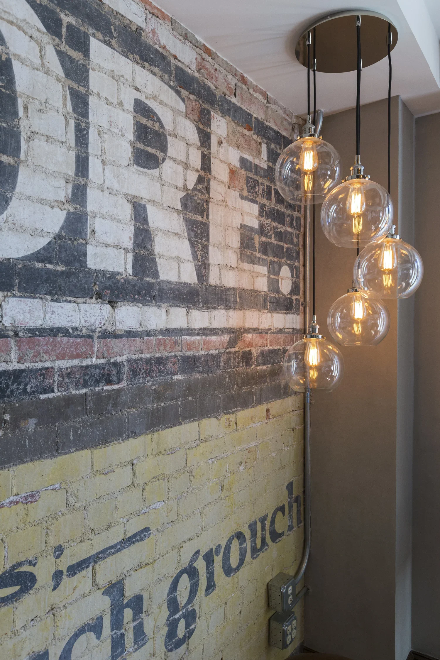 Interior with an exposed brick wall featuring faded painted text, a hanging light fixture with multiple glass globe bulbs, and electrical outlets along the wall.