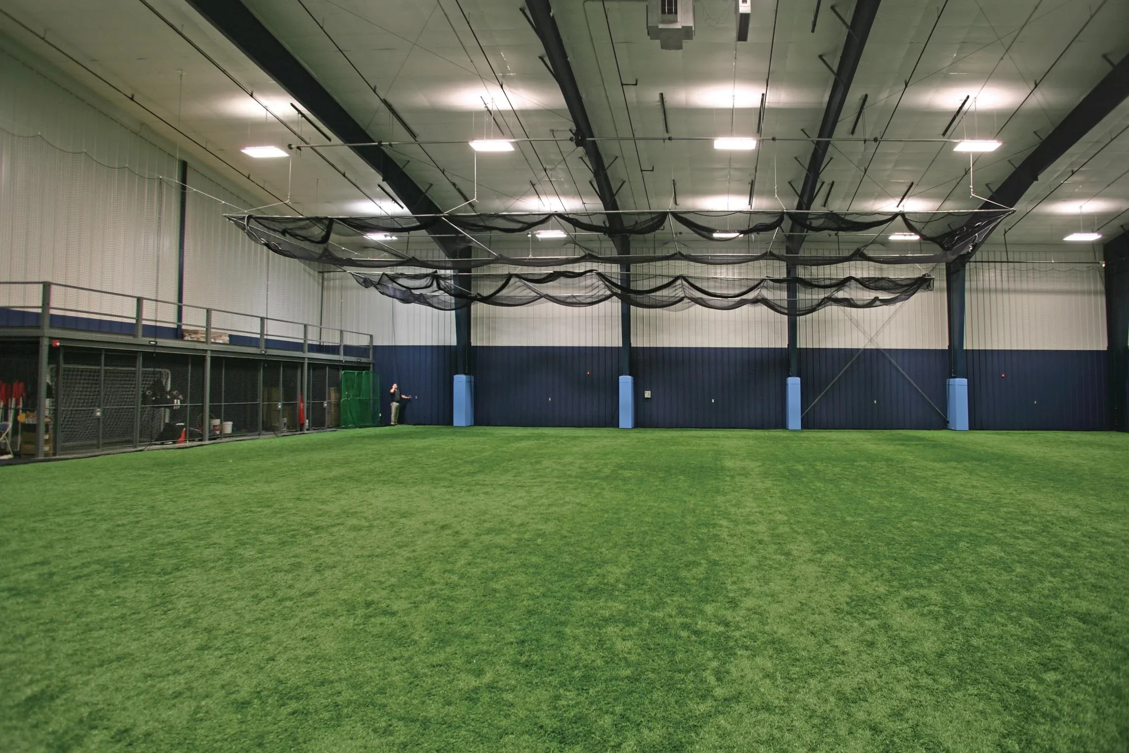 Indoor sports facility with artificial turf field, high ceiling, and large black netting hanging from the ceiling for training or practice.