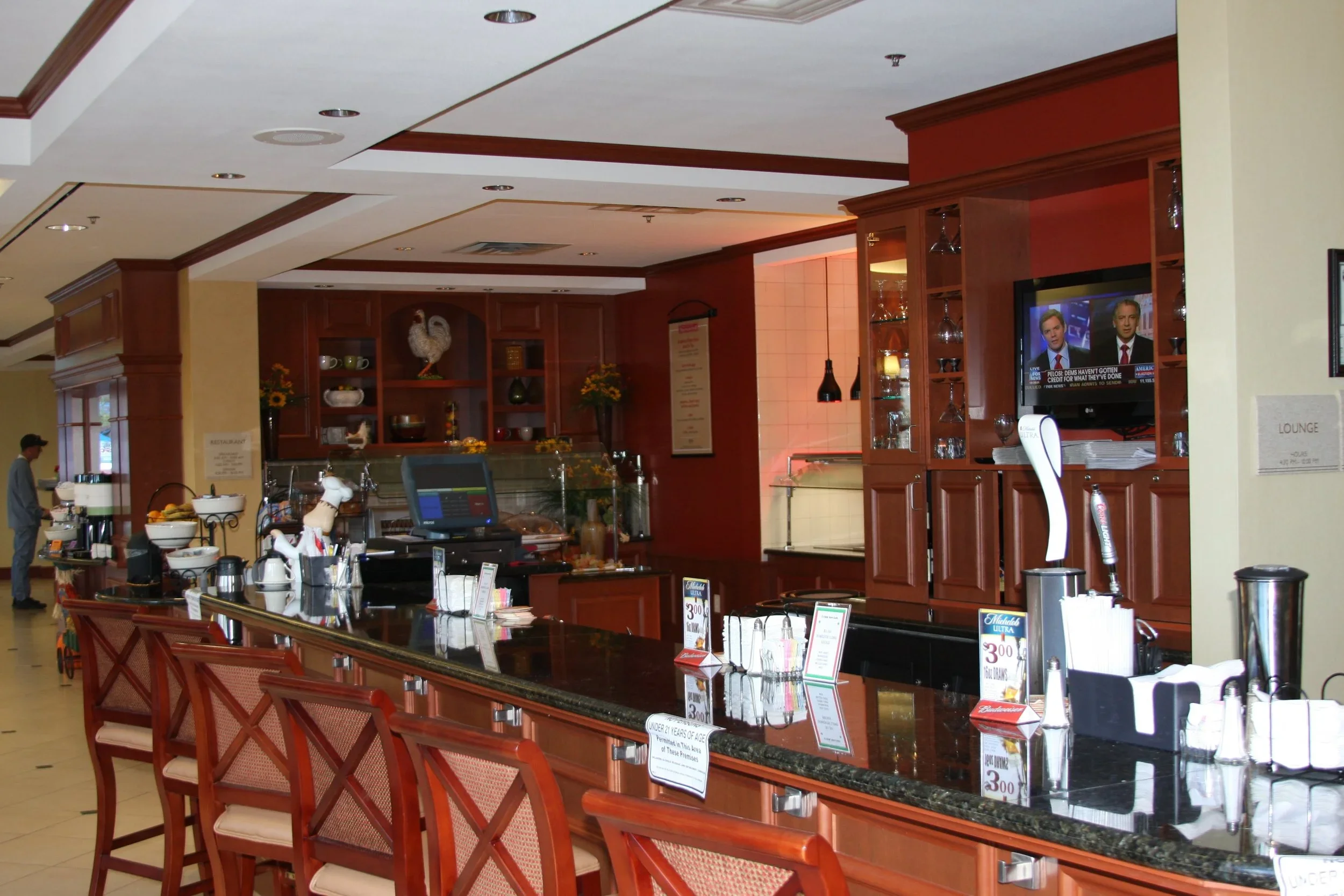 A hotel bar with a long black granite counter, wooden chairs, a mounted TV, and various beverages and equipment on the counter, with a warm wood-paneled cabinetry background.
