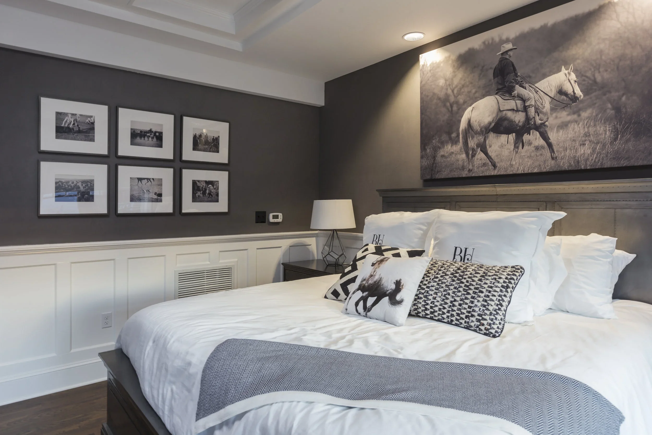A bedroom with a large bed adorned with white and patterned pillows, a dark wood nightstand with a white lamp, and black and white wall art featuring a cowboy on a horse, with additional framed photos on a dark gray wall.