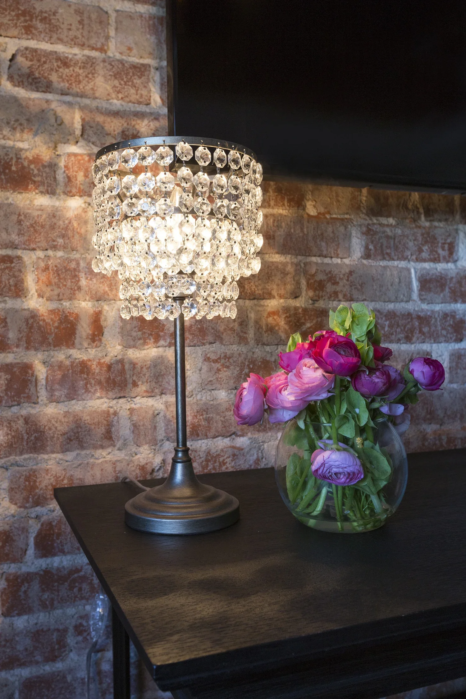 A table lamp with a shade made of hanging crystal beads and a round metal base, placed on a dark wooden surface. Next to it is a clear glass vase with pink and purple peony flowers and green leaves, against a exposed brick wall background.