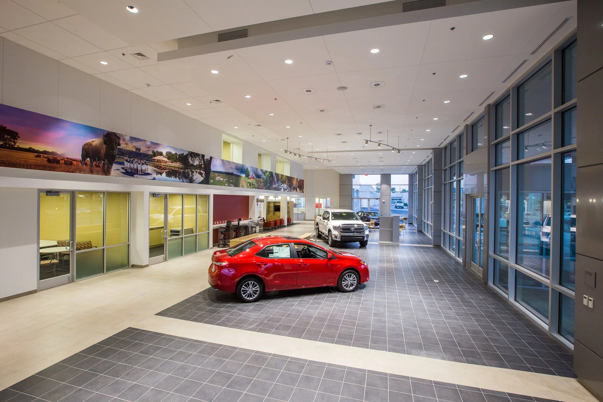 An indoor car dealership showroom with a red car in foreground, several other cars in background, and large windows along the right side.