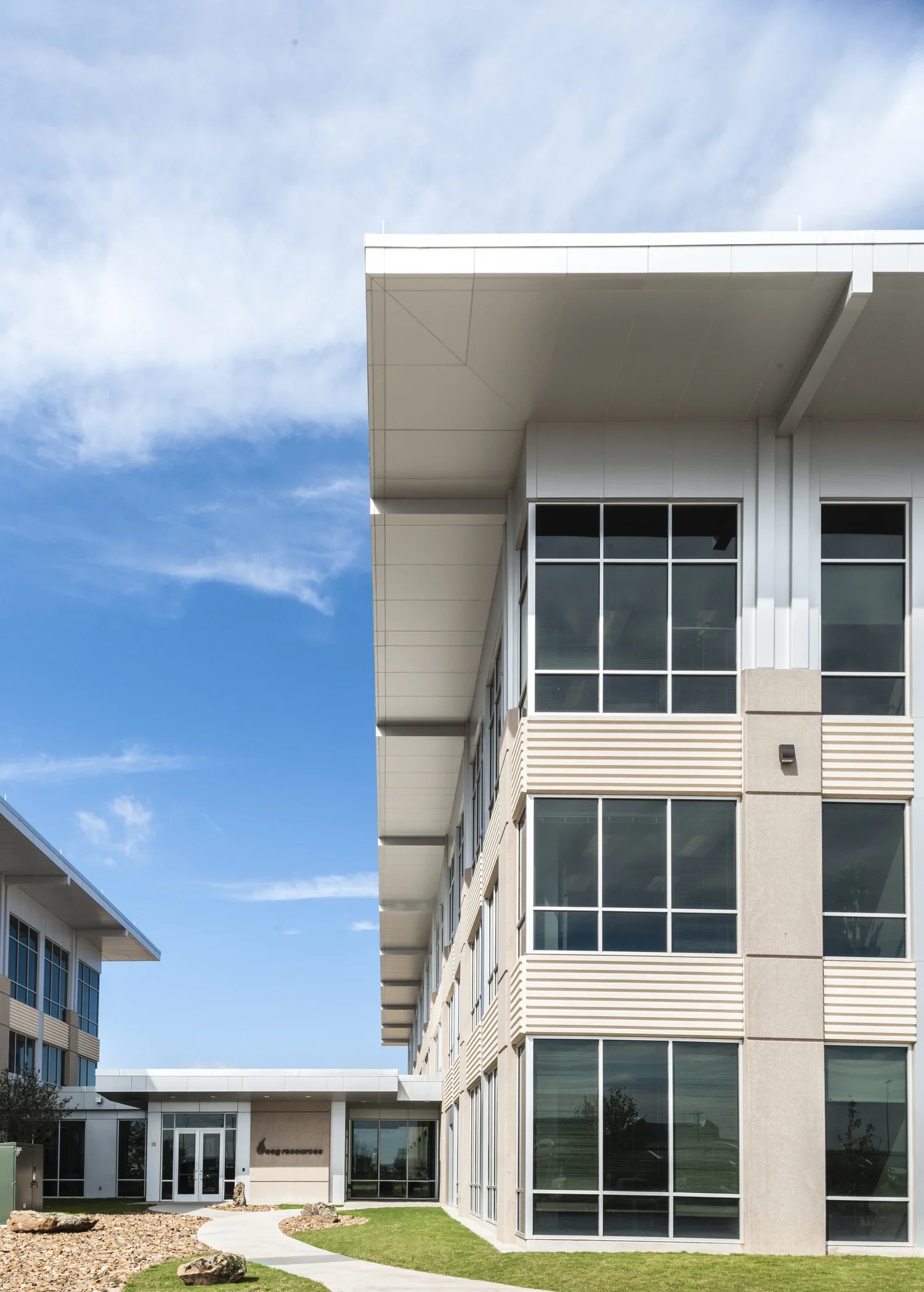 Modern multi-story building with large glass windows, a flat roof, and a landscaped front yard with rocks and grass.