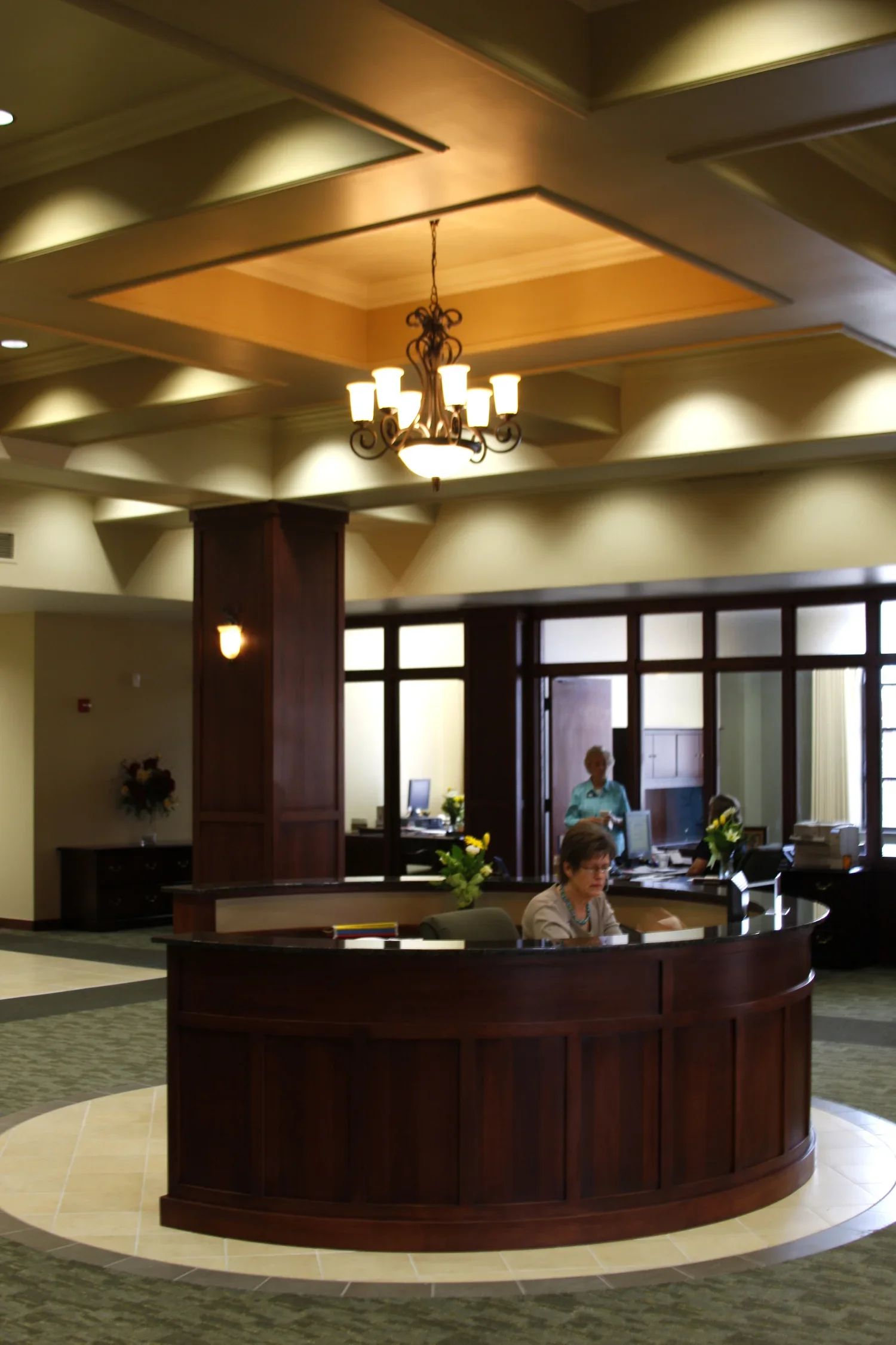 Hotel lobby reception area with a curved wooden desk, two women working behind the desk, and another woman sitting at the desk with flowers on it. There is a chandelier hanging from the ceiling and windows in the background.