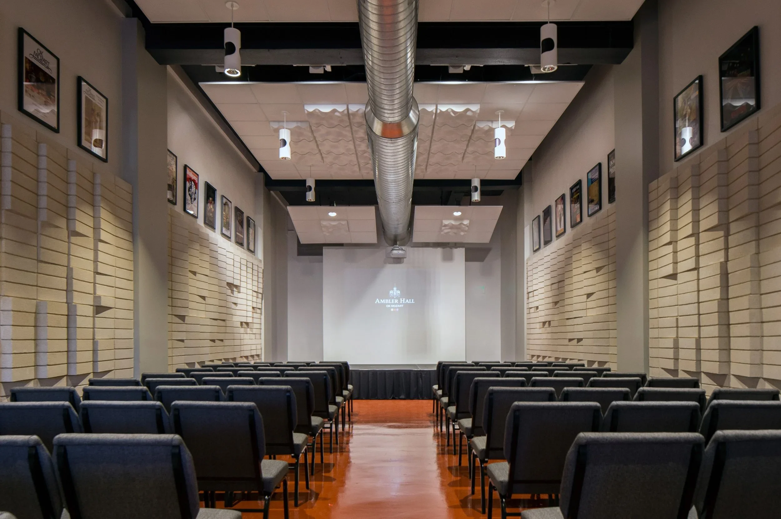 Empty auditorium with rows of black chairs facing a stage with a large screen displaying 'Ambler Hall' and 'OK Mozart' at the front. The ceiling features black and white panels with hanging light fixtures, and the walls are decorated with framed pict